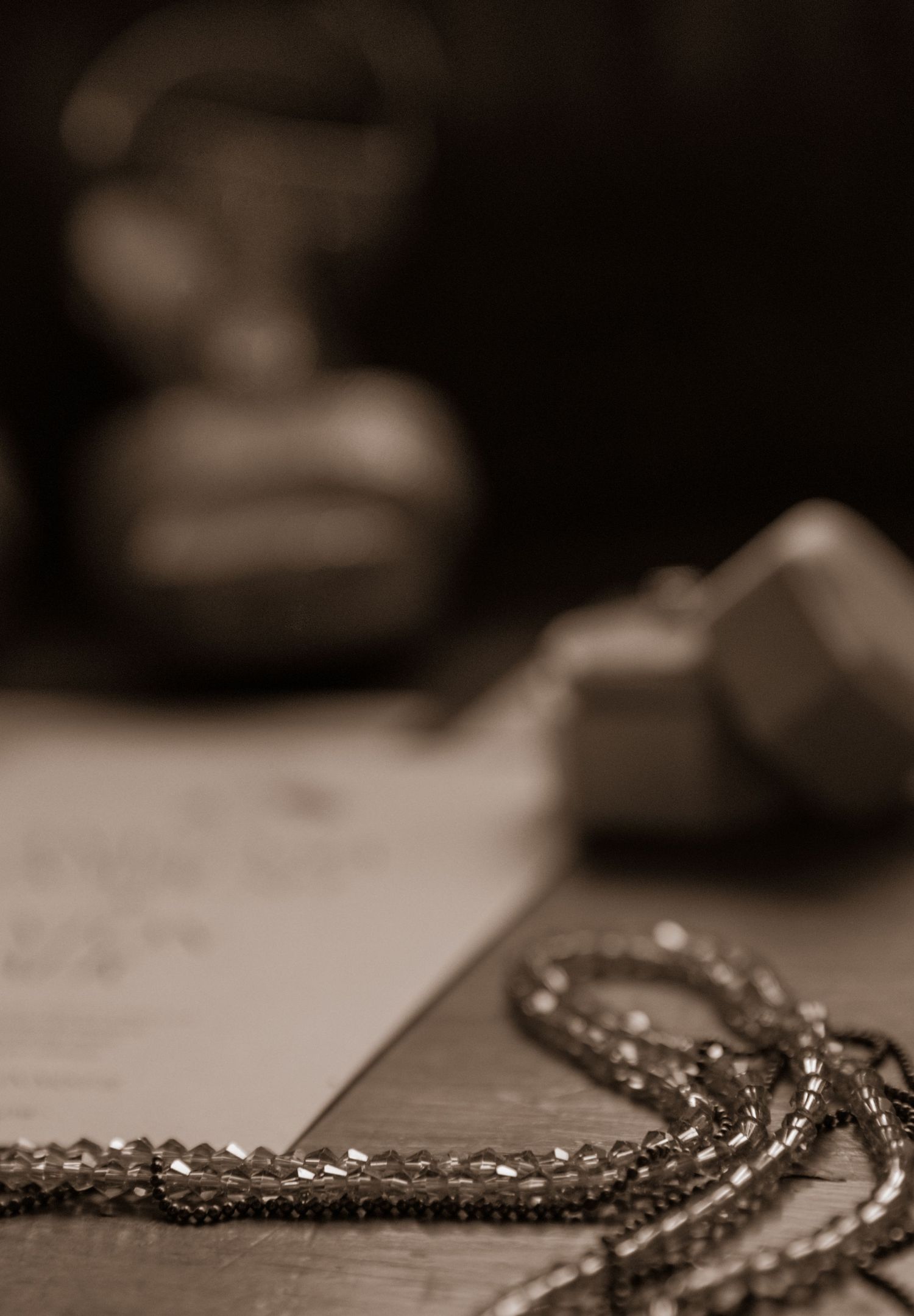 A delicate pearl necklace lies beside a blurred document in sepia tones.