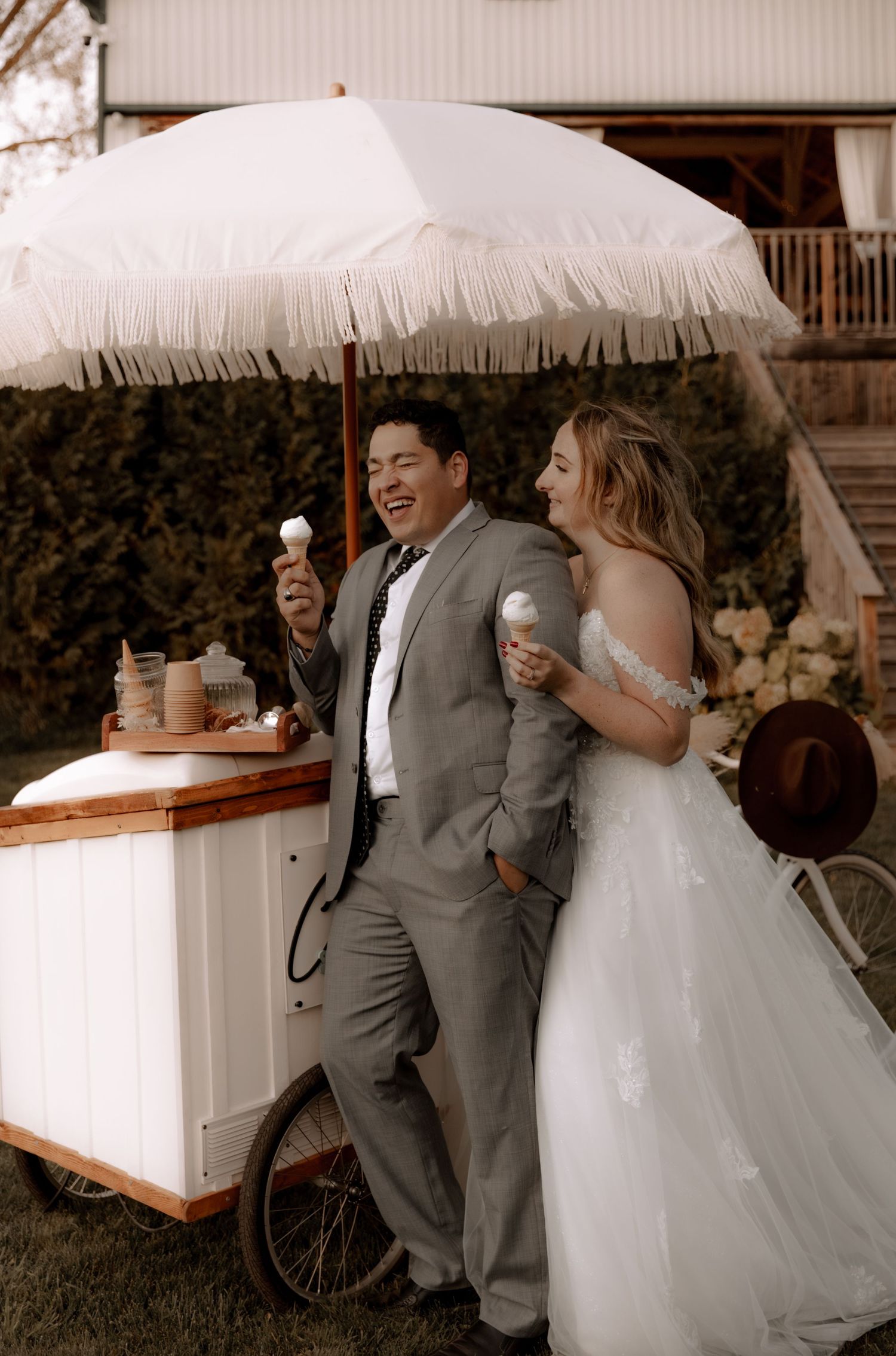 Newlyweds share ice cream under a white fringe umbrella next to a vintage cart.