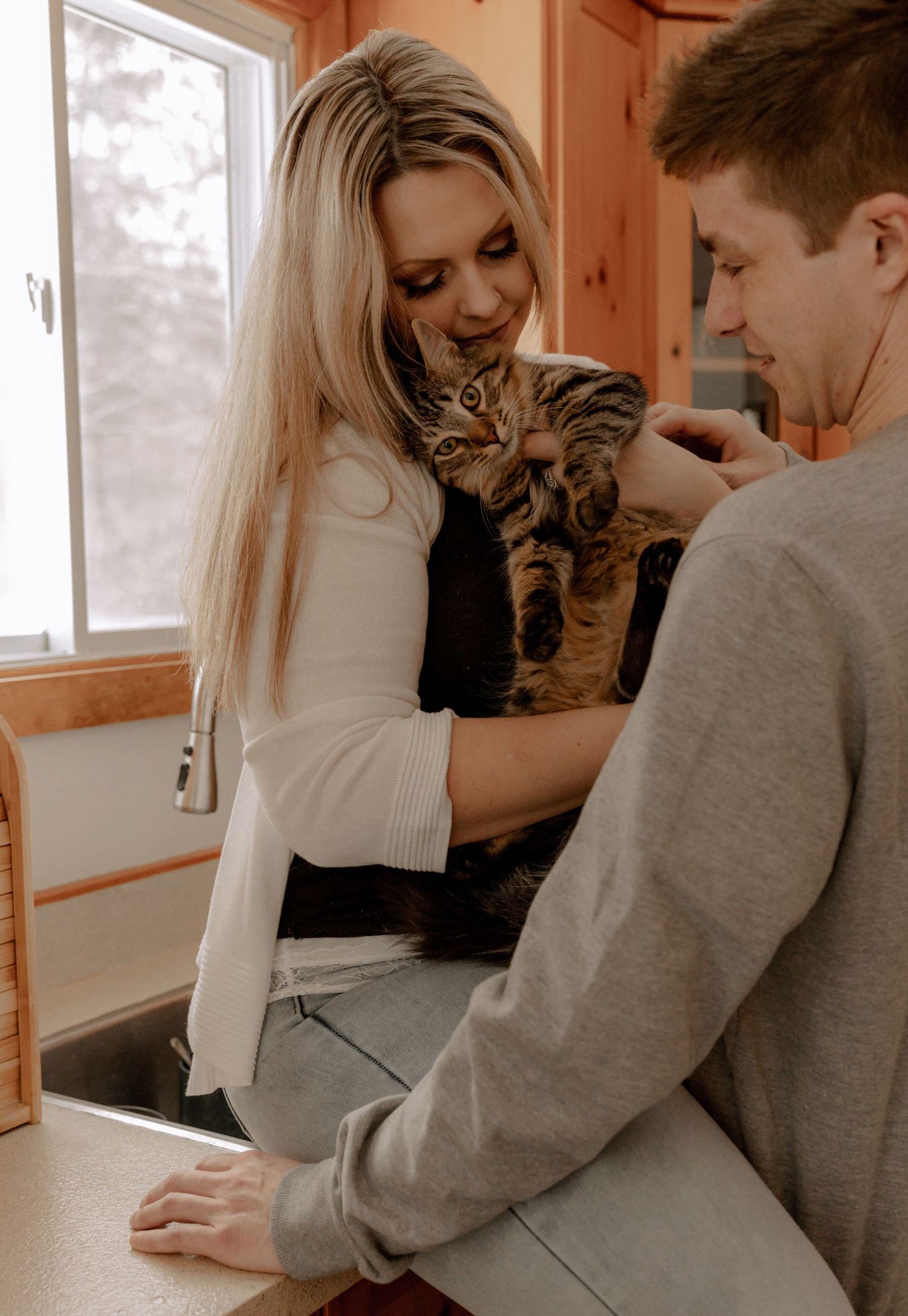 A couple shares a tender moment while holding their tabby cat in a warm, wooden-paneled room by a window.