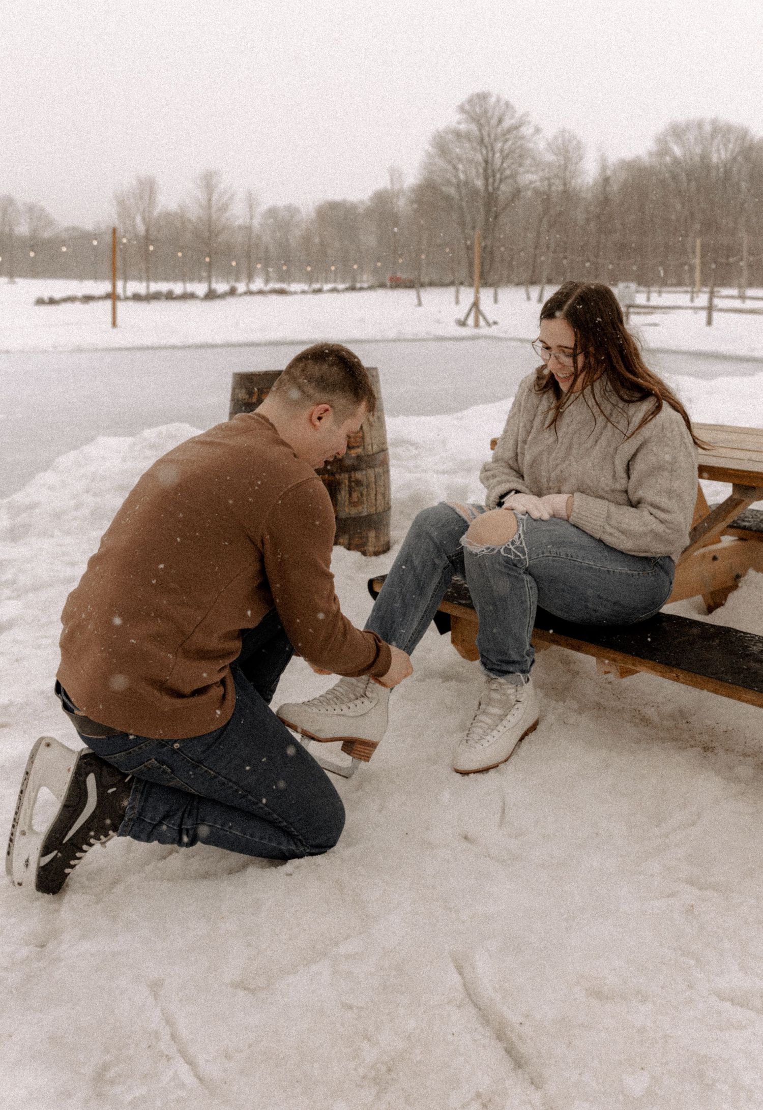 A winter scene shows two people by a snow-covered bench as one helps the other put on ice skates.