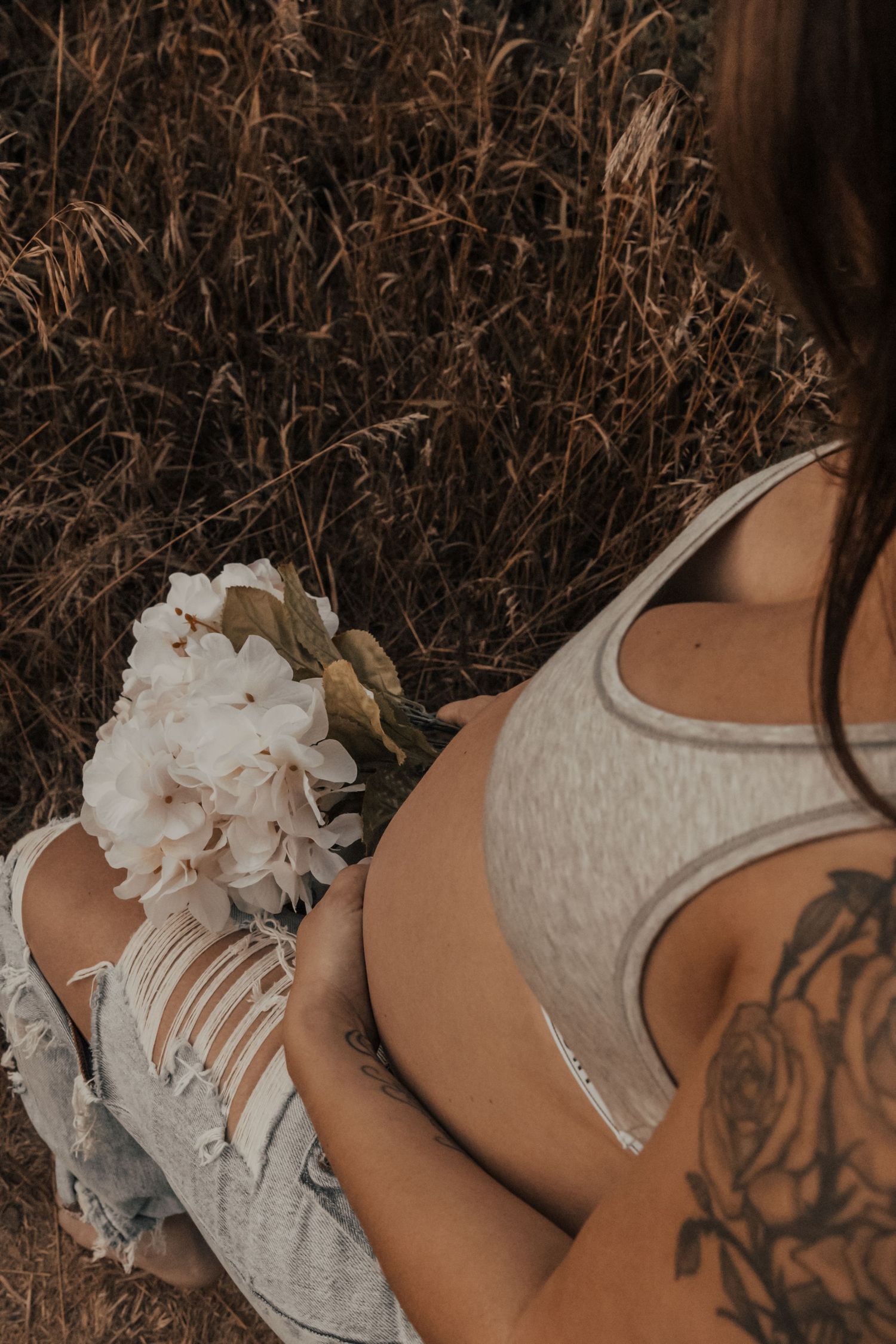 A cropped artsy photo showing delicate white flowers and a tattoo detail against dried tall grass in sepia tones.