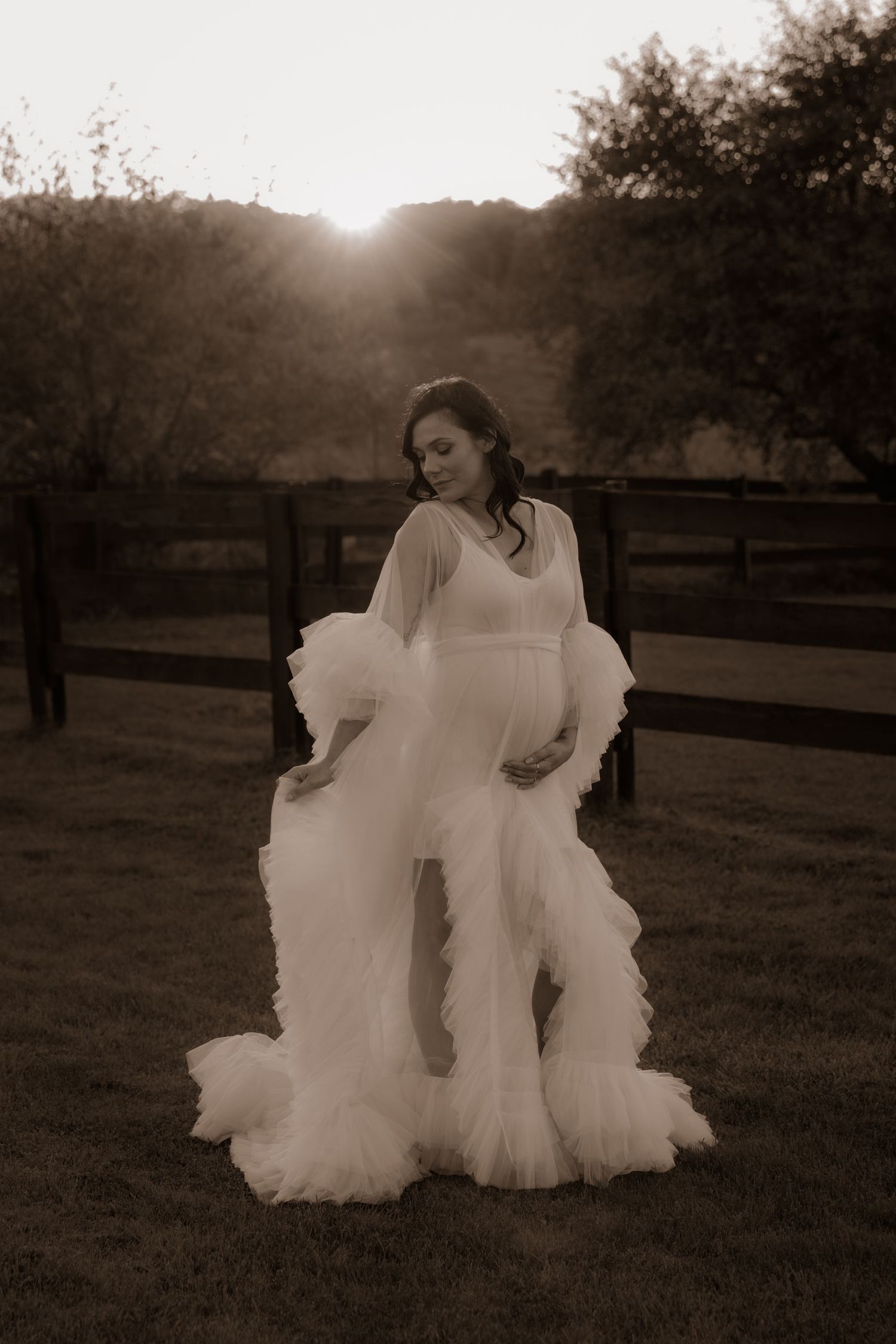 A sepia-toned photo of a flowing white dress twirling in a backlit outdoor sunset setting with wooden fencing.