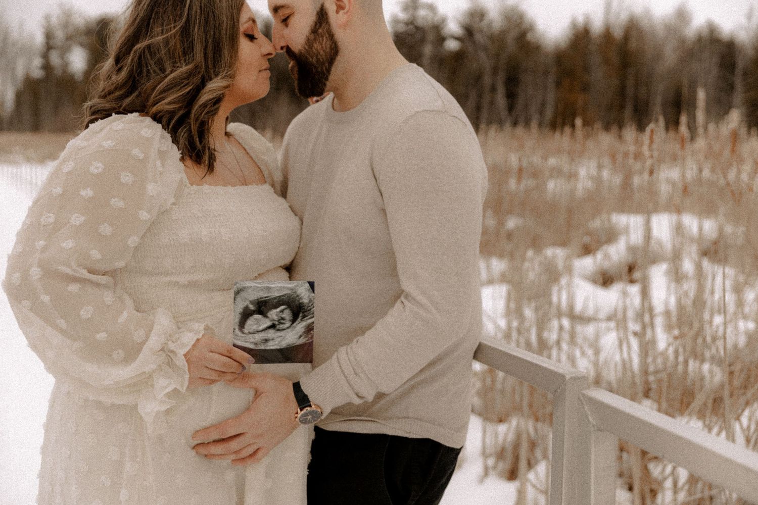 Two people in winter attire share an intimate moment while holding a sonogram photo in a snowy field landscape.