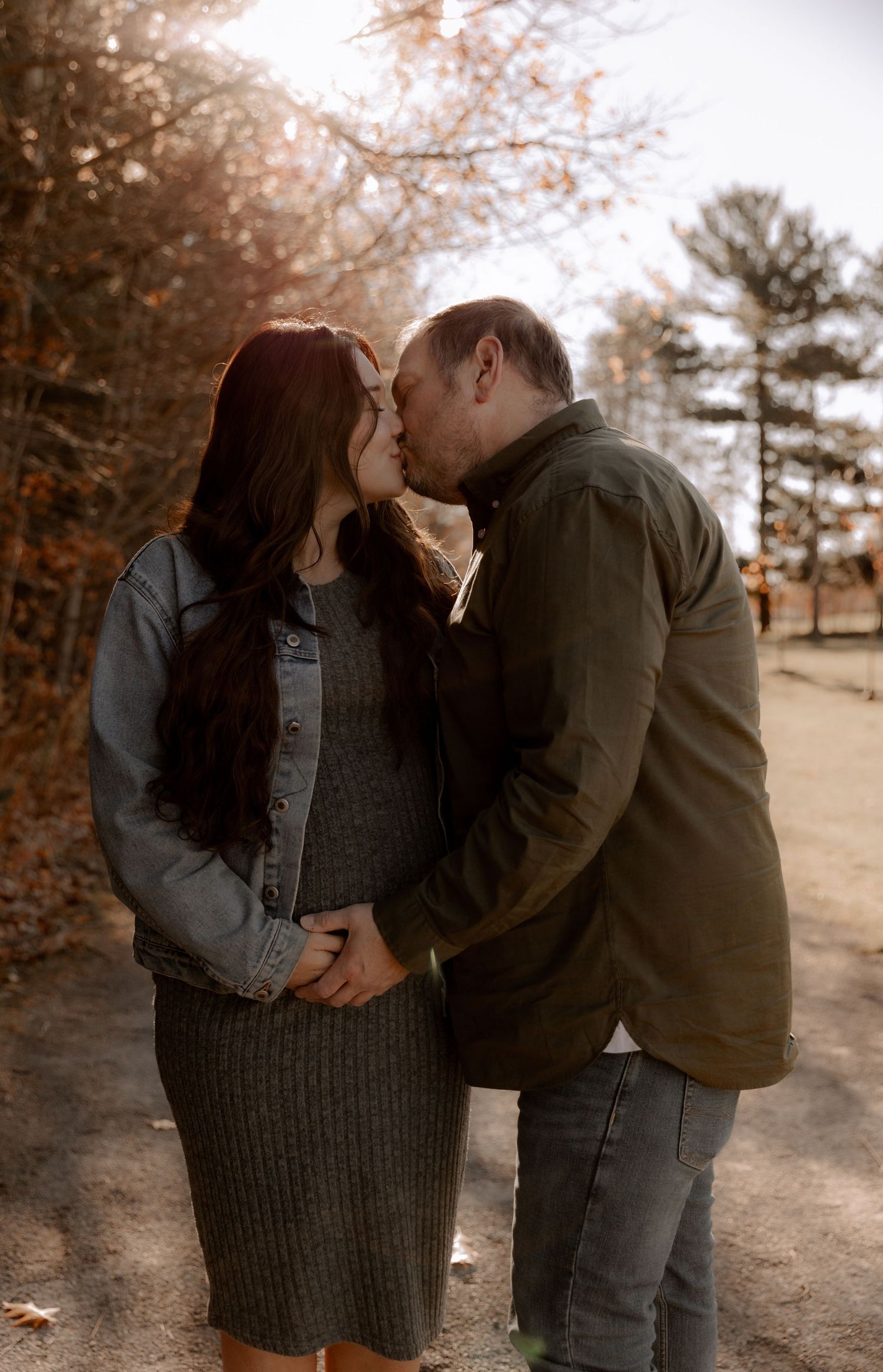 A couple shares a romantic kiss on a path surrounded by autumn trees and fallen leaves in warm golden light.