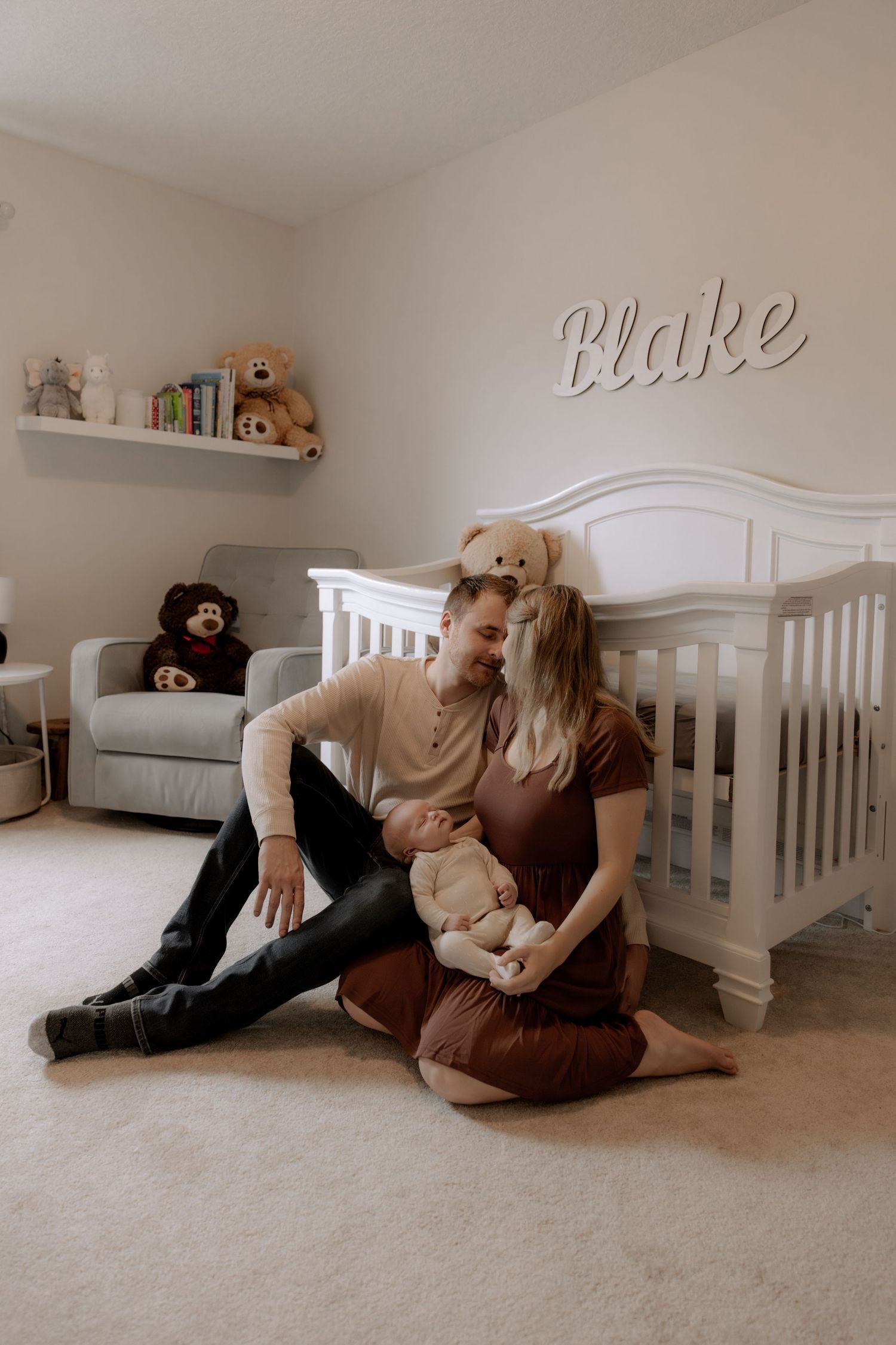 Baby nursery with white crib, gray armchair, floating shelf with toys, and 'Blake' name decor on neutral walls and carpet.