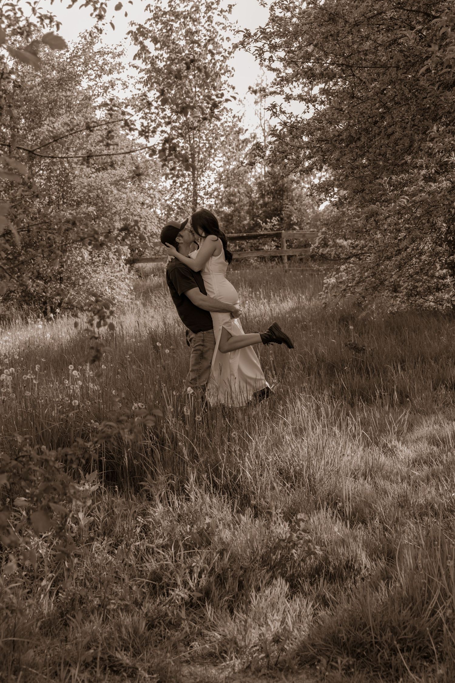 A romantic black and white photograph shows a couple embracing in a sunlit meadow surrounded by trees.