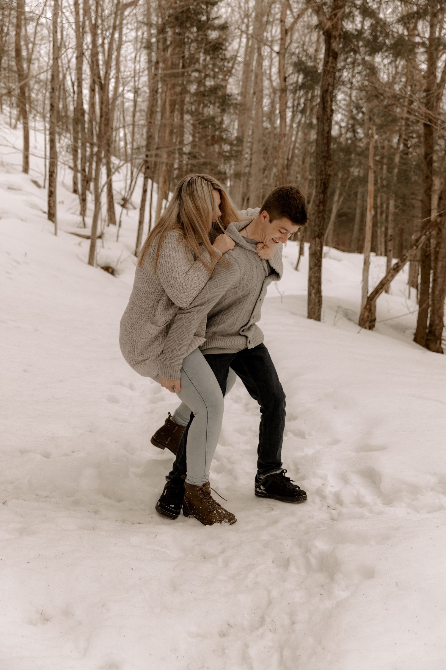 A couple in winter clothing embrace and laugh together on a snowy forest path during a romantic winter photoshoot.