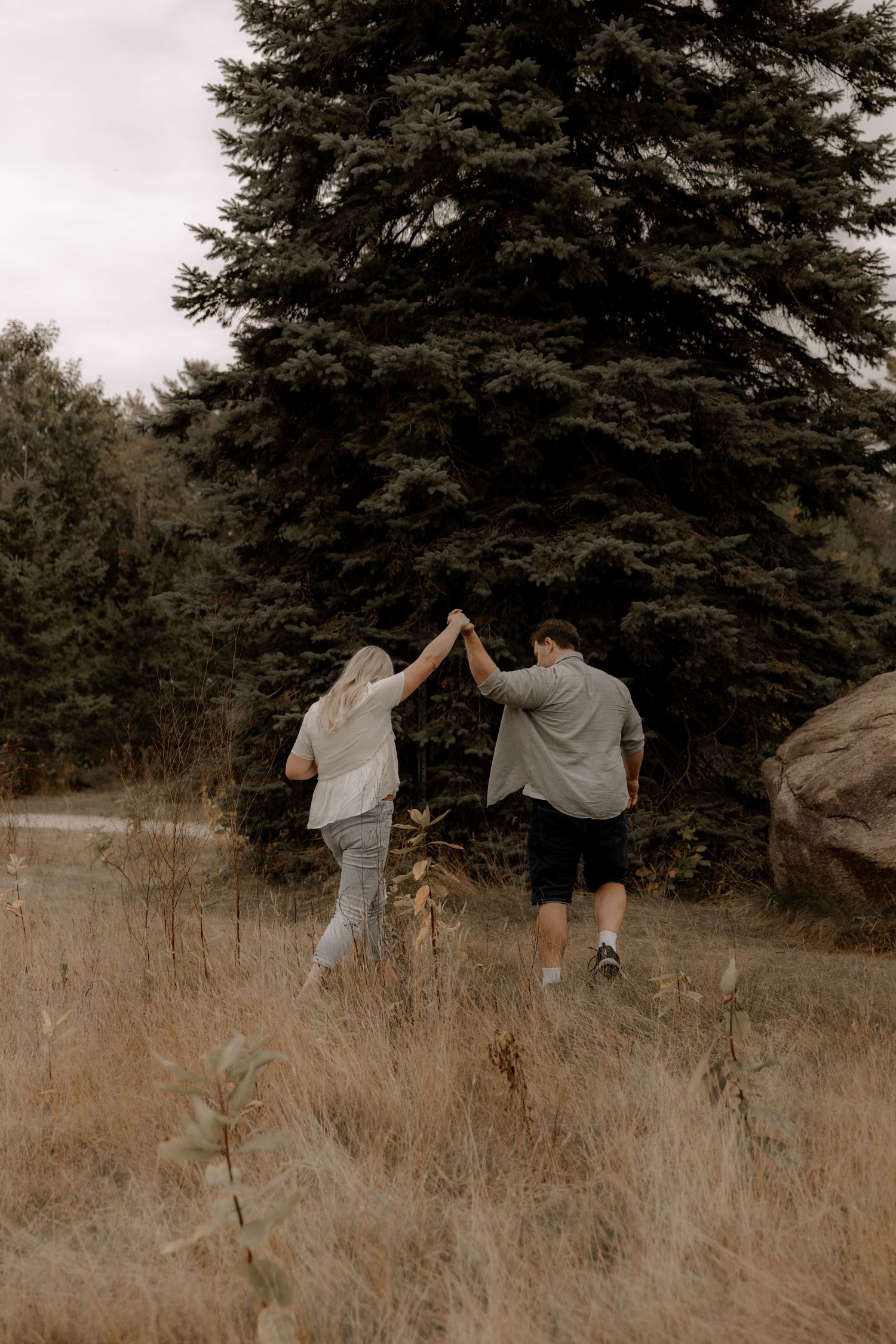 A playful couple holding hands while walking through a field with a tall evergreen tree in the background.