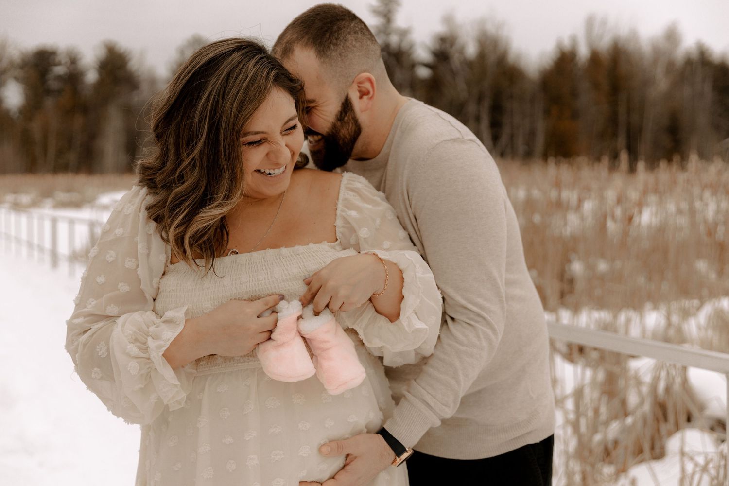 A couple holds tiny pink baby shoes in a snowy winter field during an intimate photoshoot.