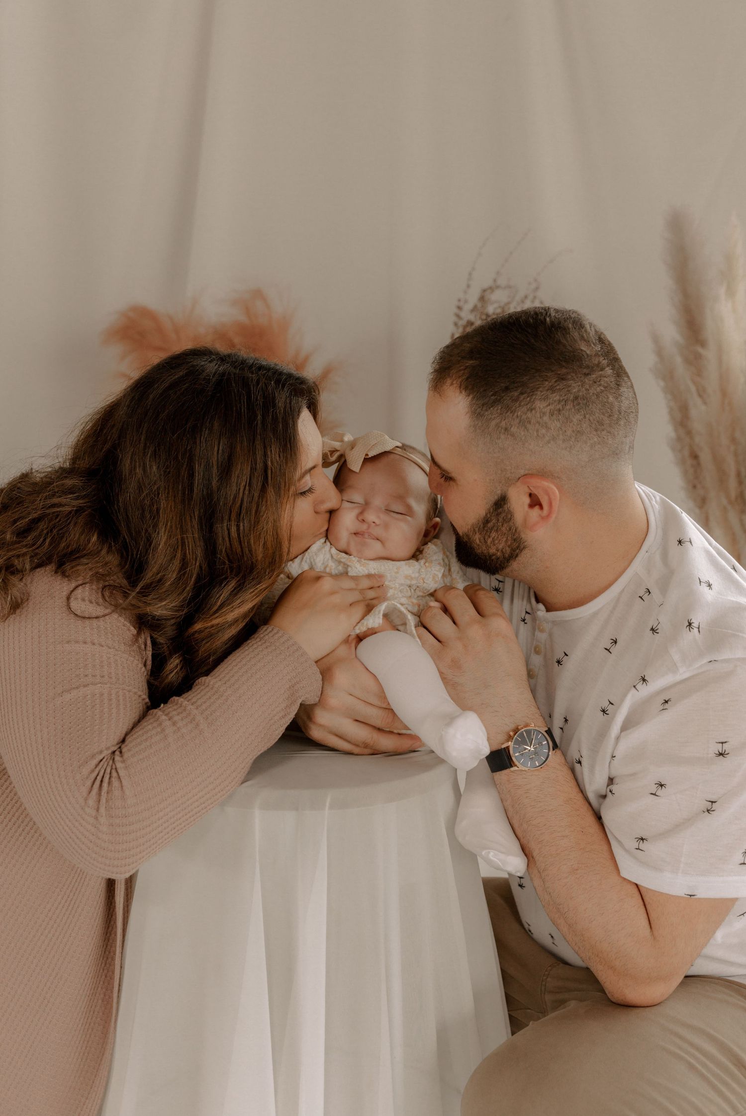 Parents share tender moment with newborn baby in soft neutral studio setting against beige backdrop.