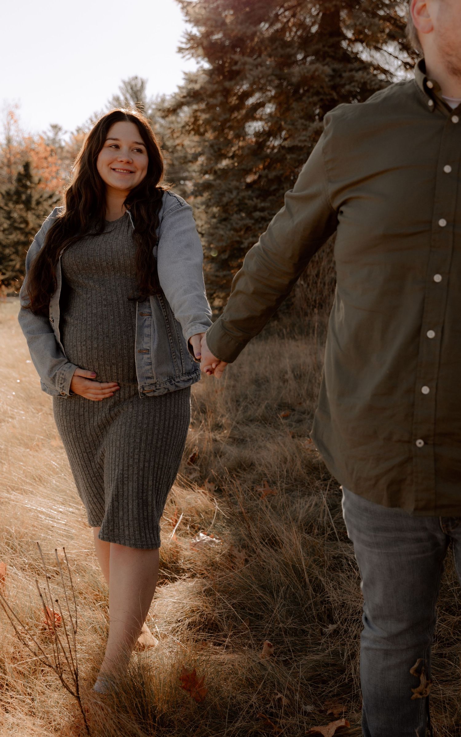 A couple holds hands on an autumn path while person in grey sweater dress and denim jacket smiles warmly.