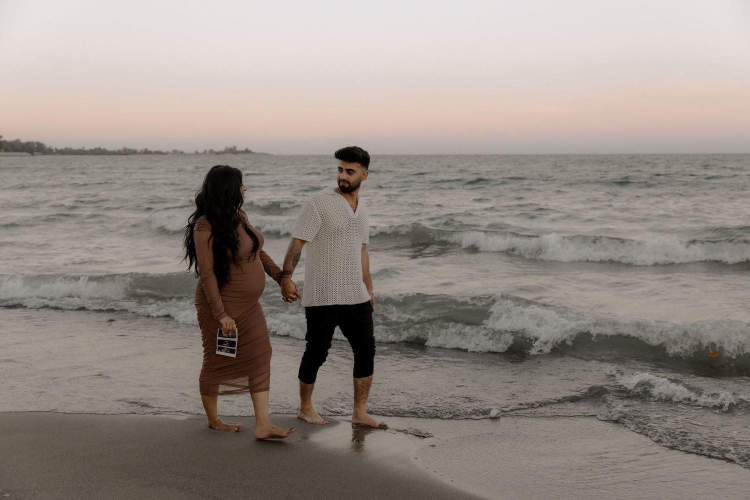 Two people standing together on a beach at sunset with waves crashing in the background.