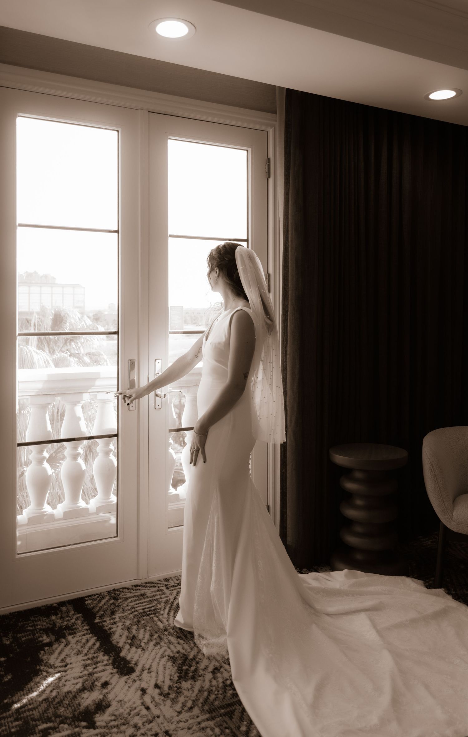 A bride in a fitted wedding dress gazes through French doors in a dreamy indoor setting.