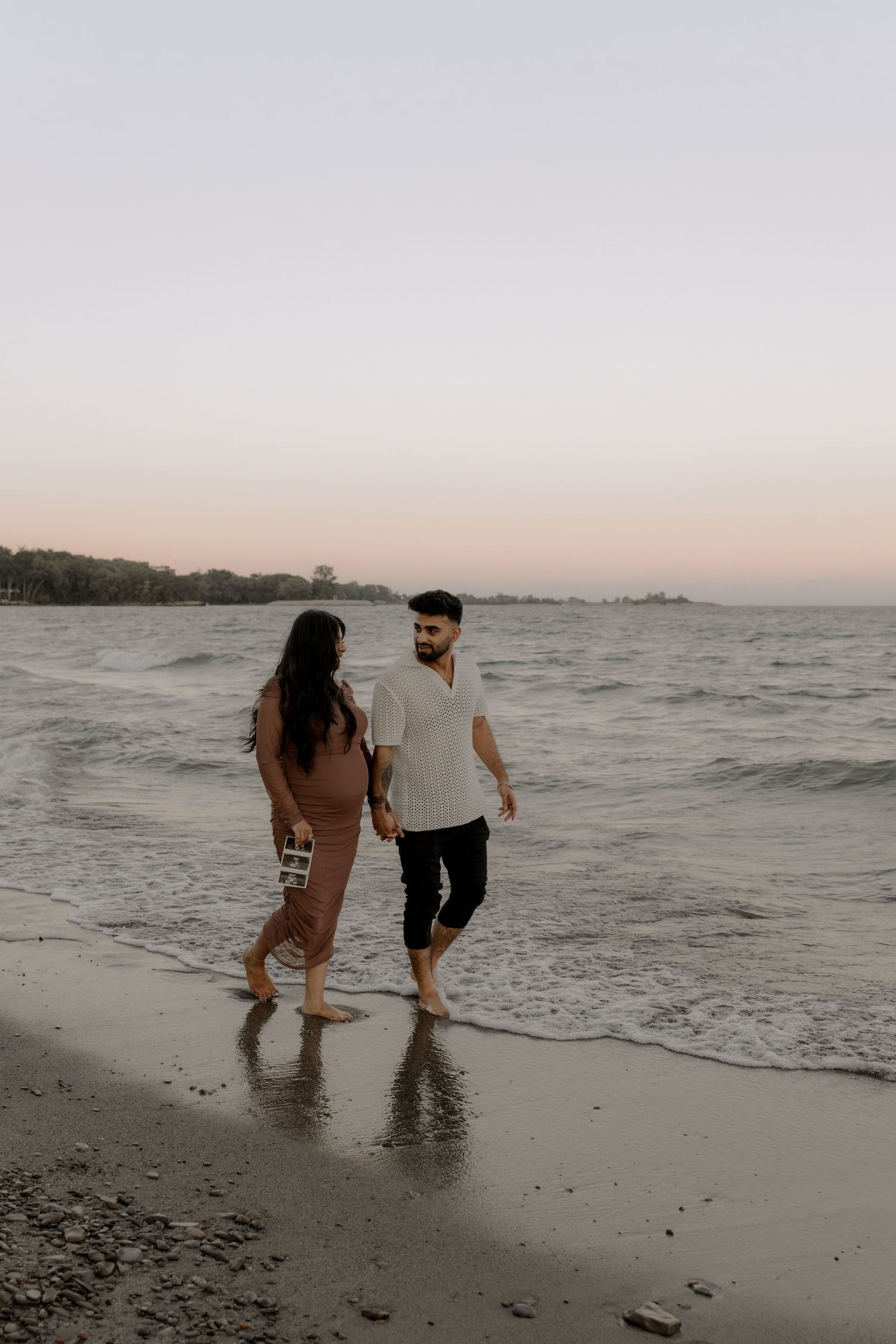 Couple enjoying romantic beach walk while holding ultrasound photos.