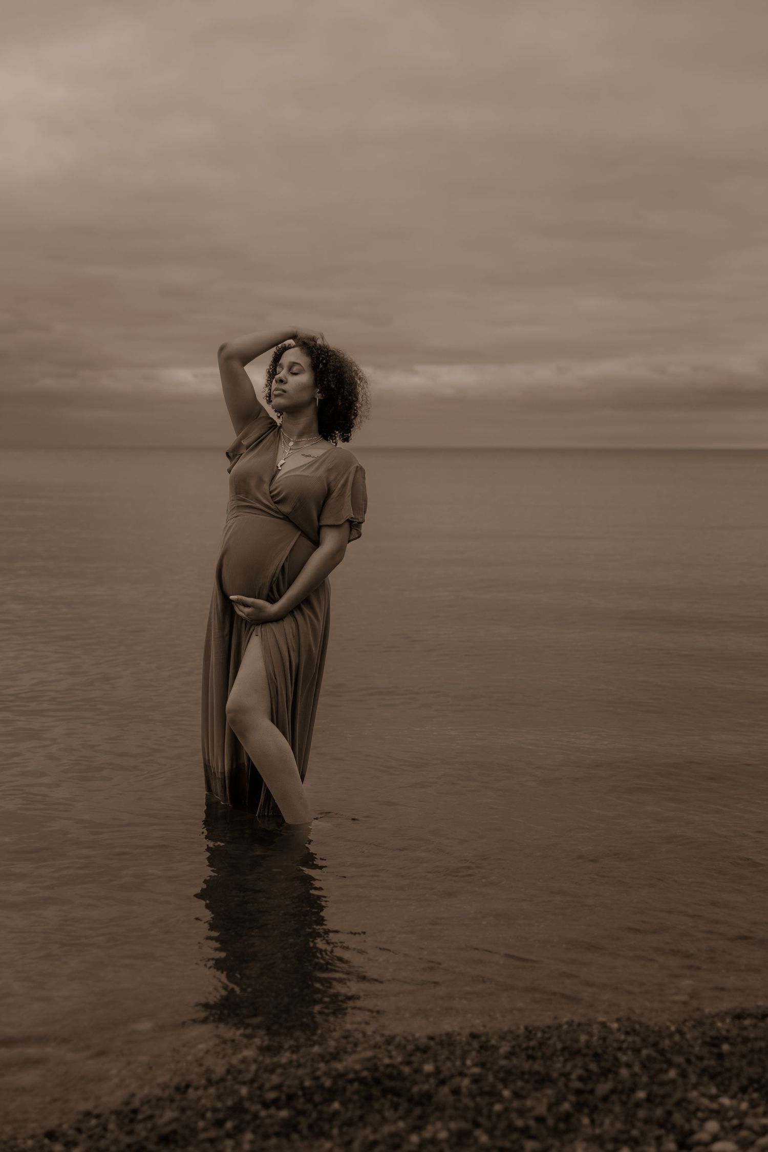 An artistic maternity portrait of a woman in a flowing dress standing in calm waters at sunset.