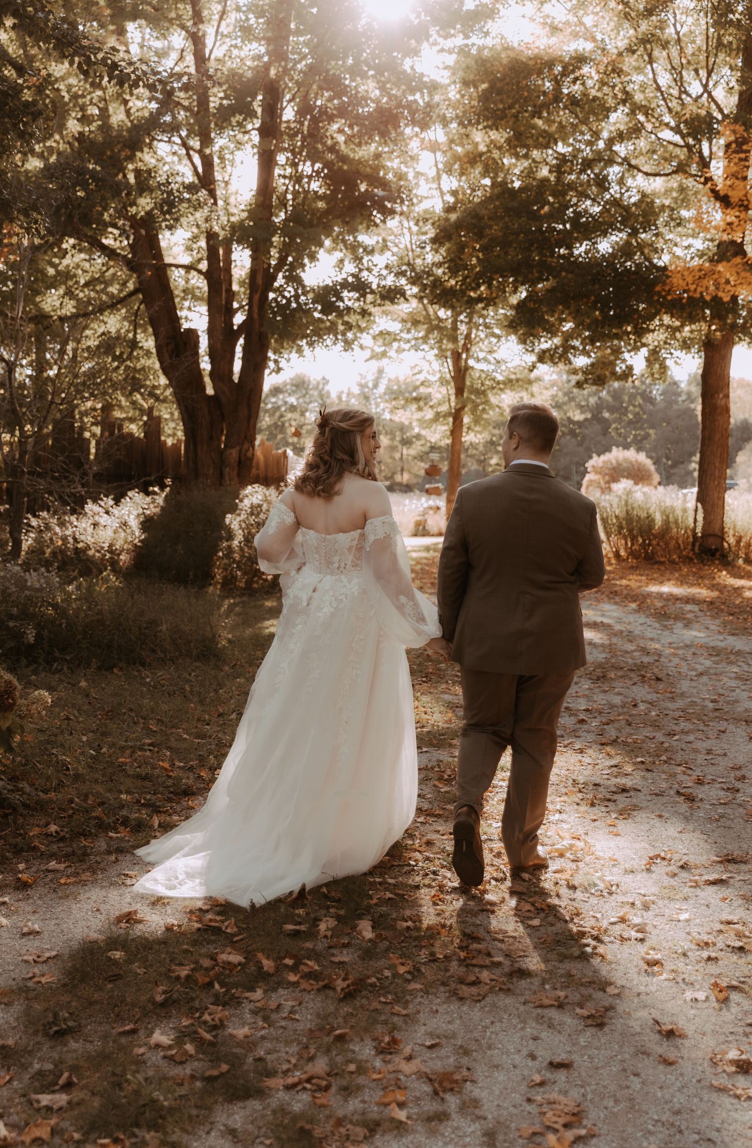 Wedding couple walking down sunlit autumn path surrounded by golden trees.