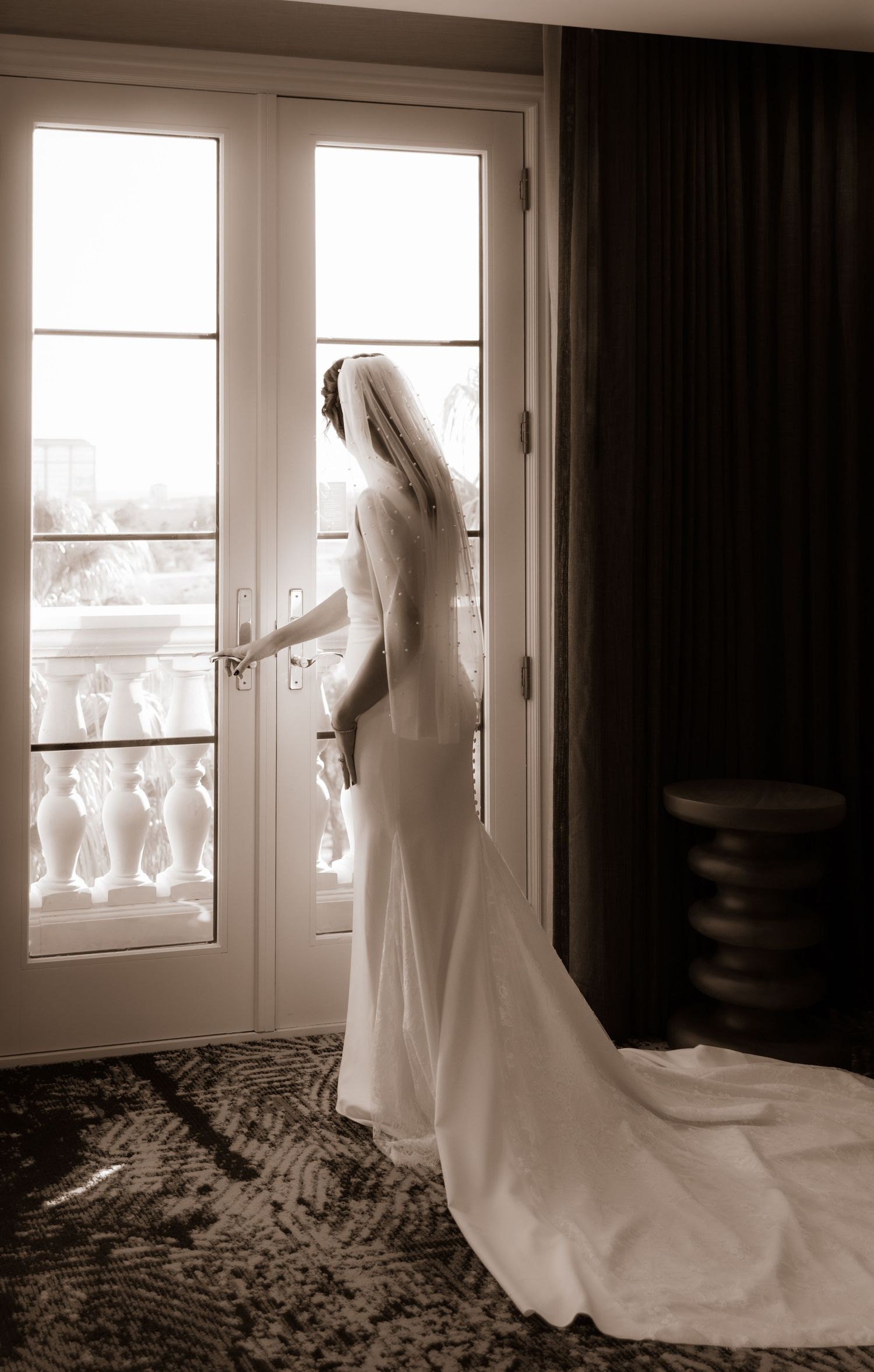 A bride in a fitted wedding gown with train stands backlit at French doors creating a dramatic silhouette.