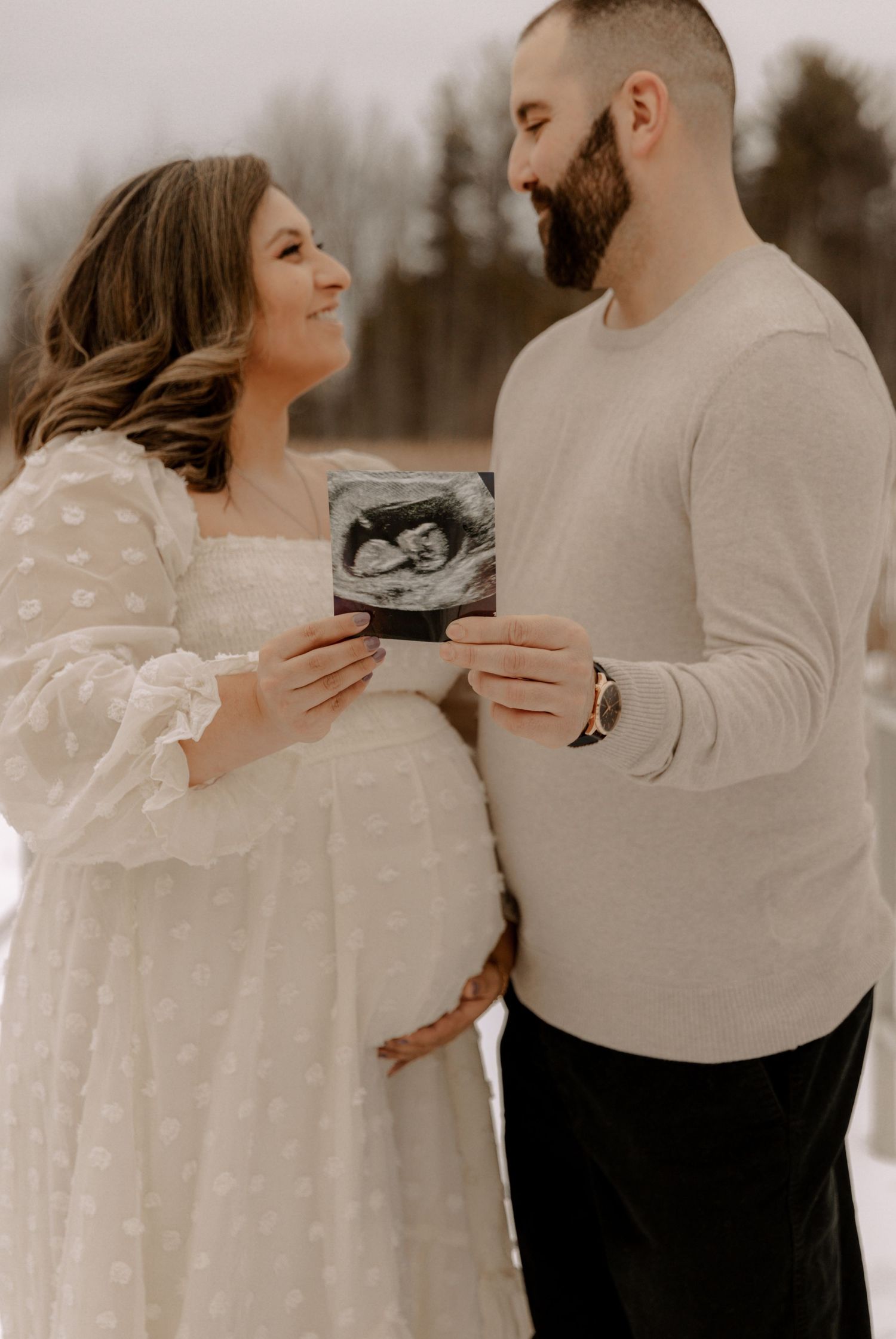 A couple in casual winter attire share a joyful moment while holding an ultrasound photo in an outdoor setting.