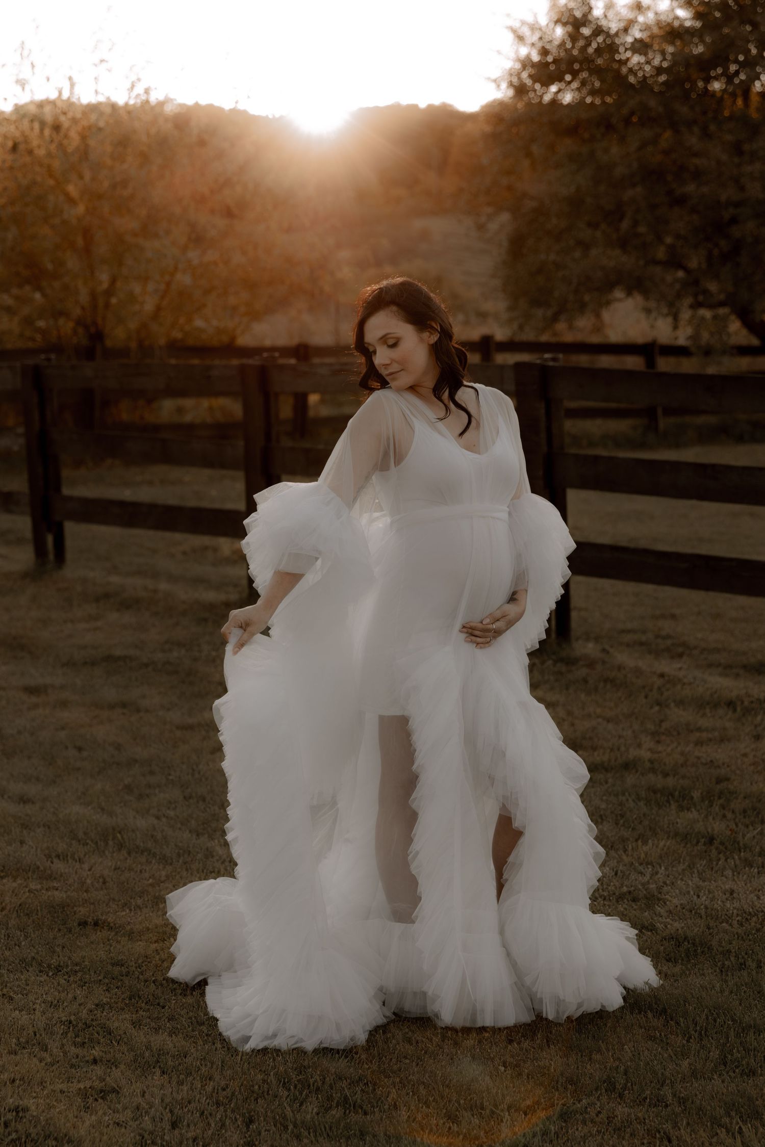 A dreamy maternity photoshoot at sunset featuring a flowing white ruffled gown in a rustic farm setting.