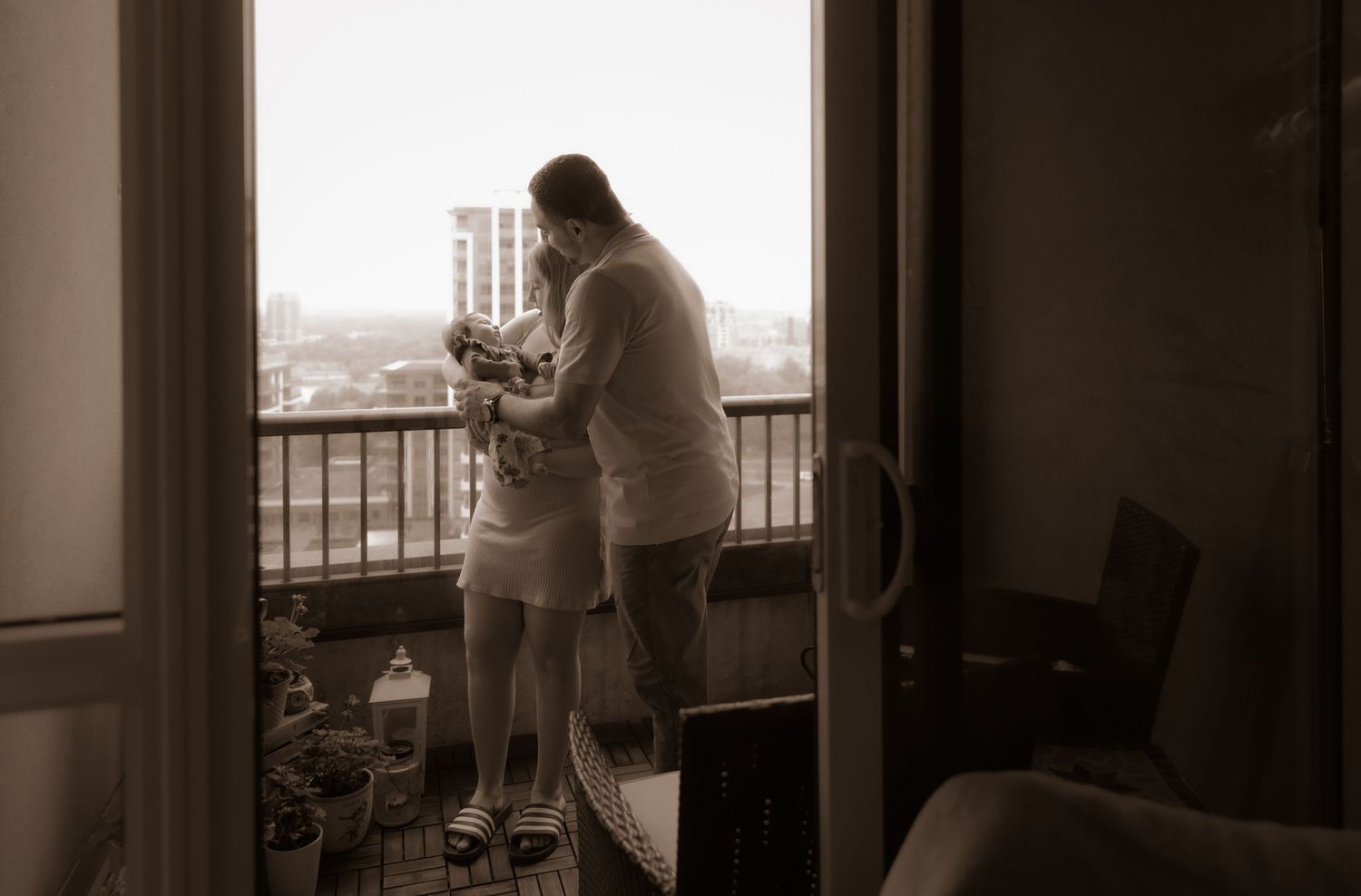 Two figures stand together on an apartment balcony overlooking a city skyline in a sepia-toned photograph.