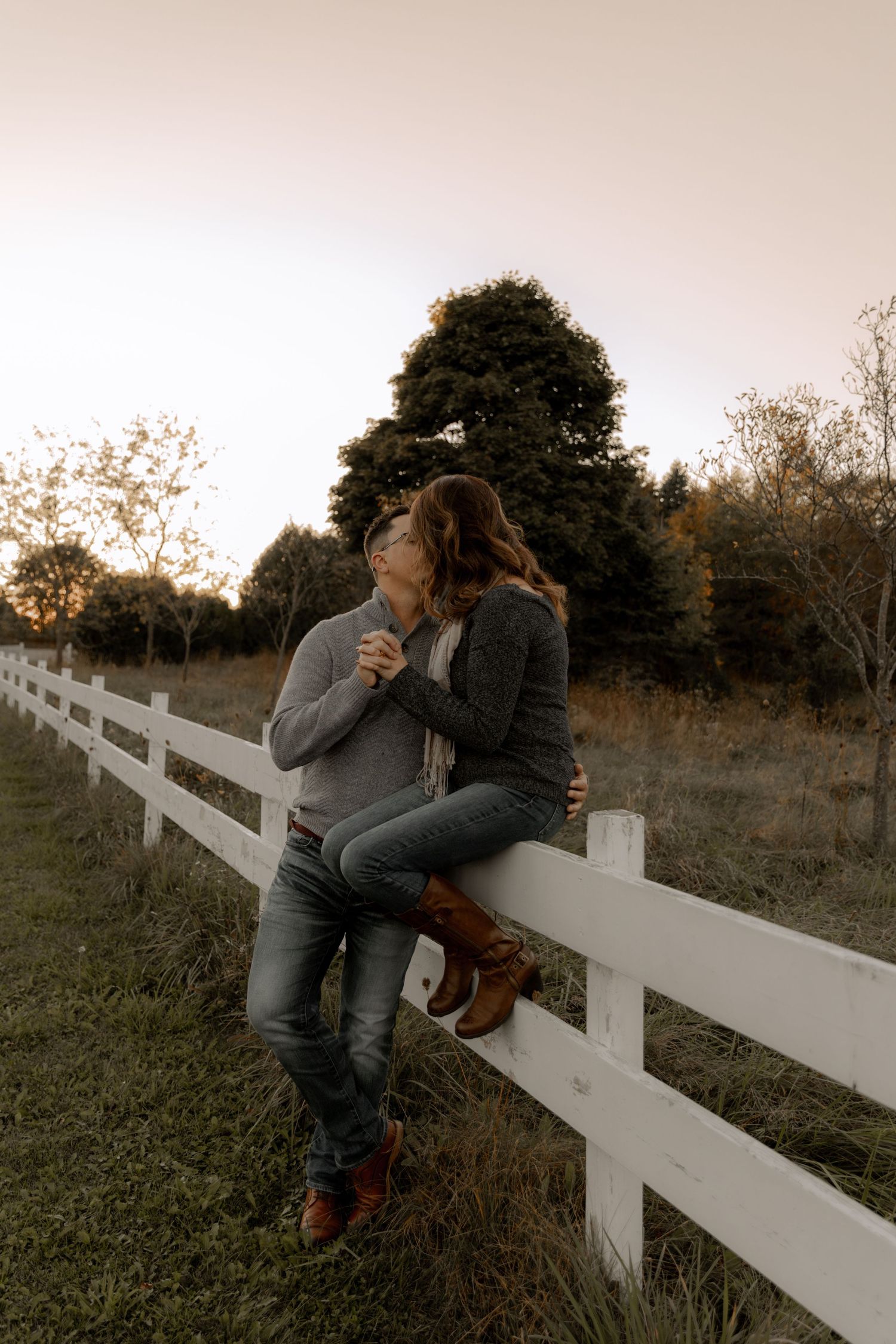 A romantic couple sitting on a white fence at sunset with fall foliage in the background.