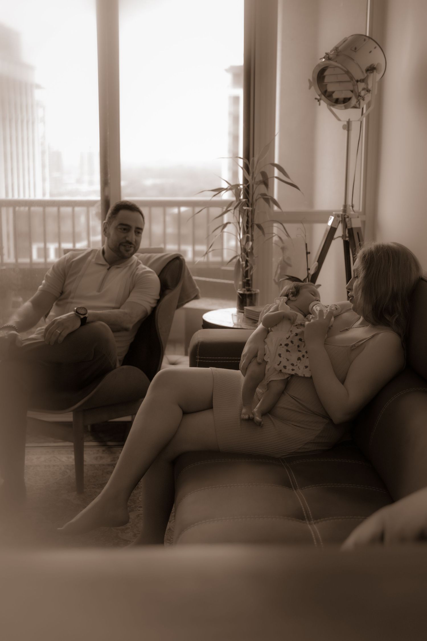 People relaxing on a couch in a living room with soft sepia tones and a potted plant visible on the balcony.