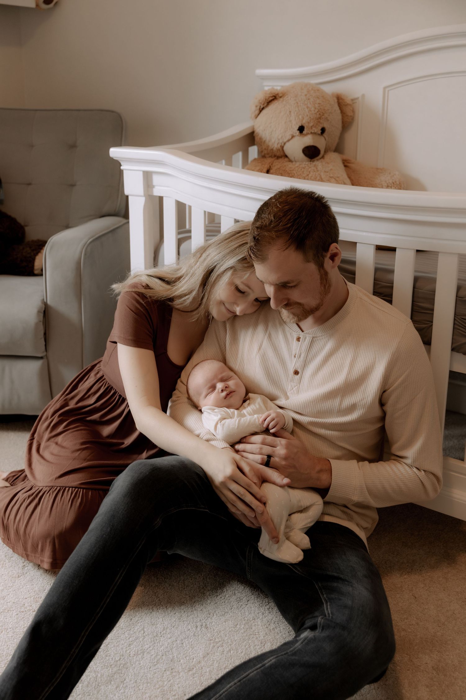 A tender moment in a nursery as parents cradle their newborn baby beside a white crib with a teddy bear watching.