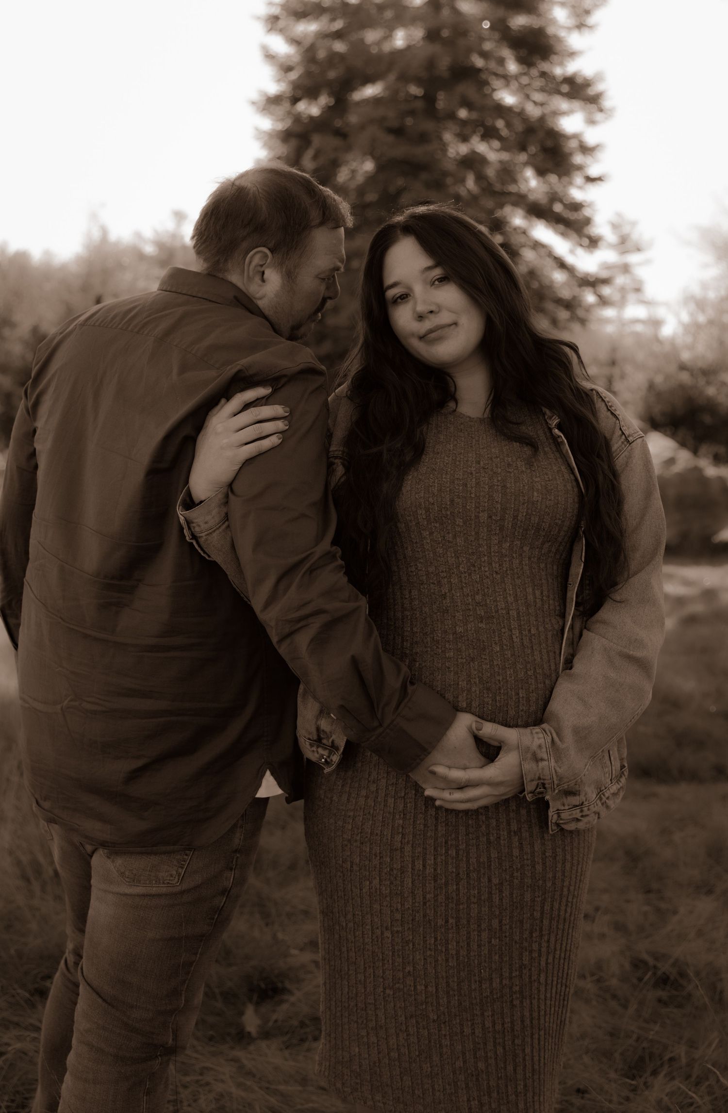 Sepia-toned outdoor romantic photo of a couple embracing, with the person wearing a brown knit dress in late autumn setting.