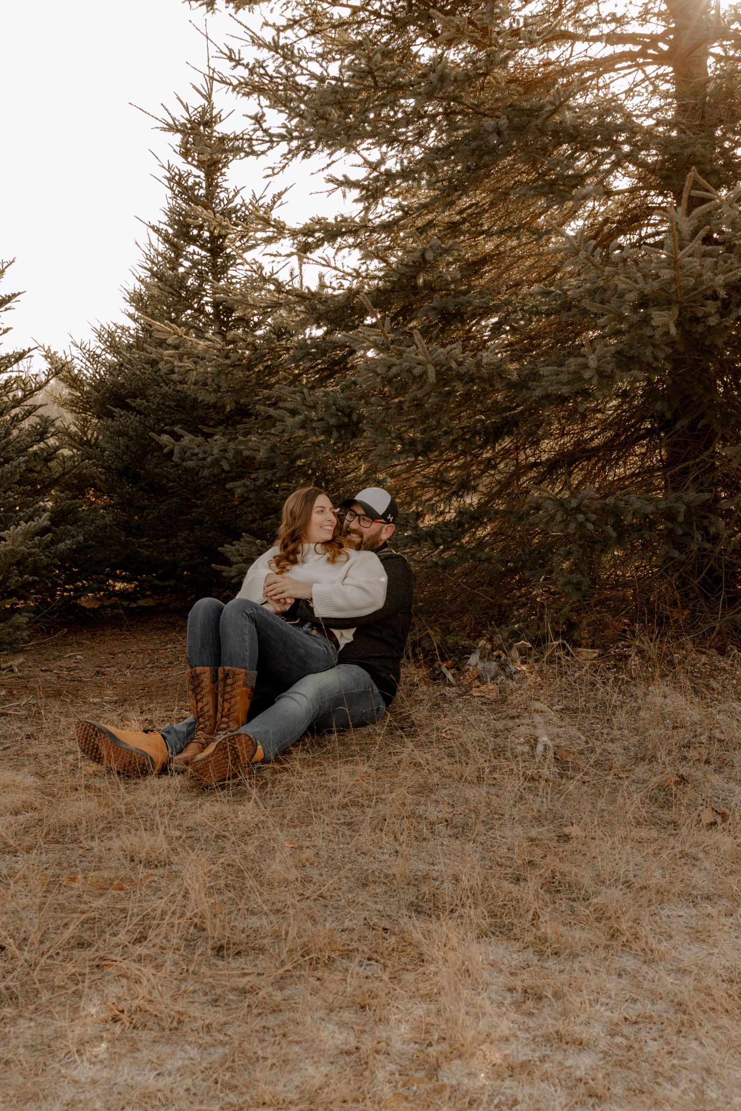 A couple sits together against evergreen trees in a rustic outdoor setting during golden hour.