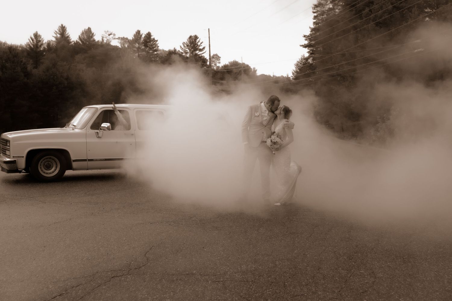 Sepia toned image capturing a couple embracing in swirling smoke near a classic vehicle.