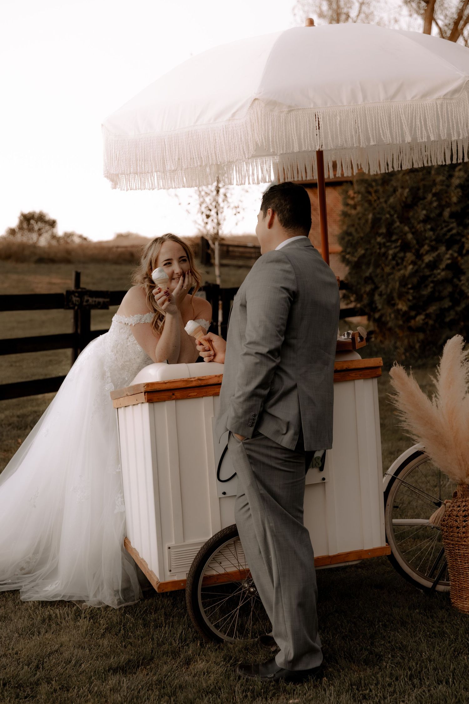 Couple enjoys ice cream from a vintage cart under a white fringed umbrella at an outdoor wedding reception.