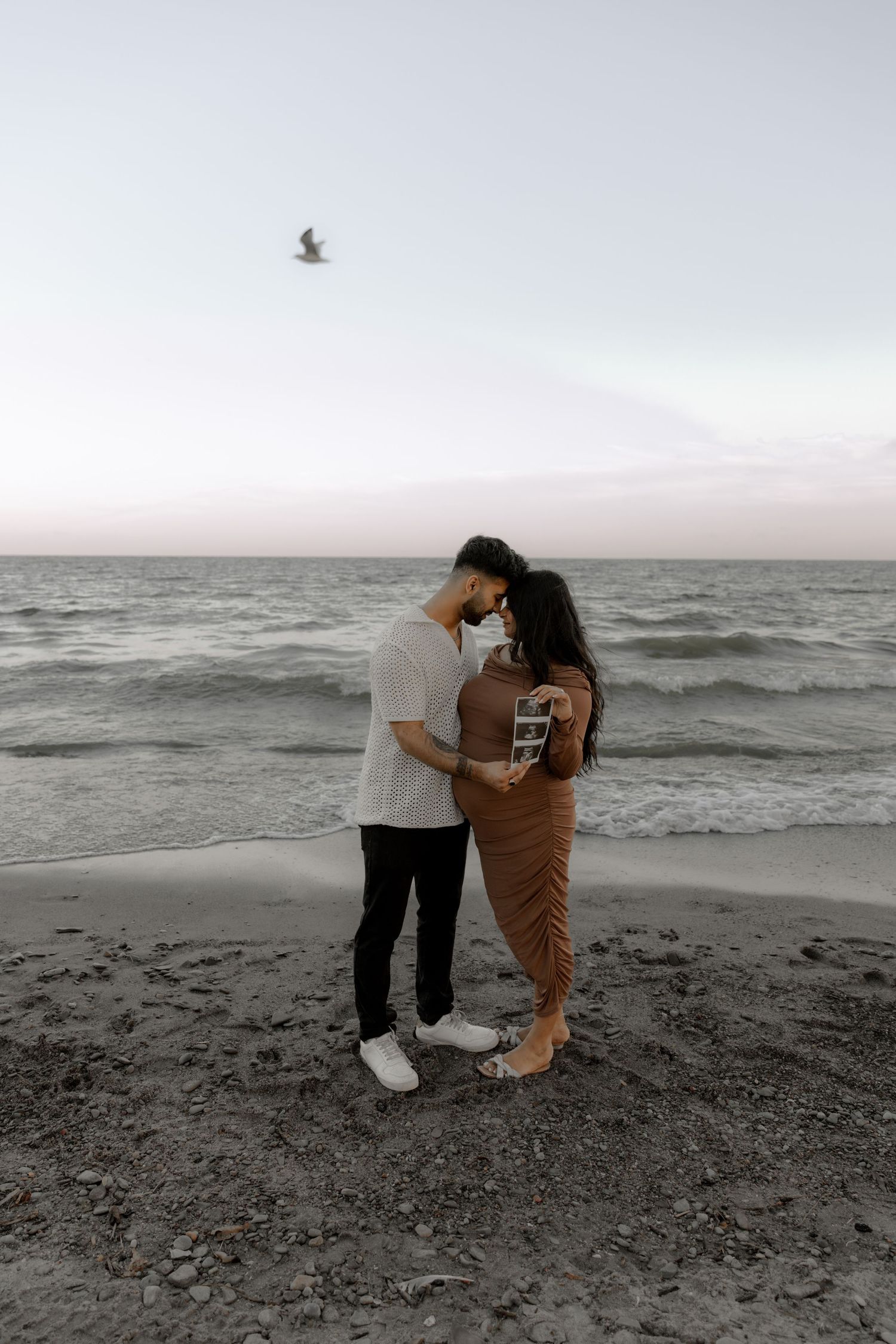 Expectant couple embraces on sandy beach while looking at ultrasound images.