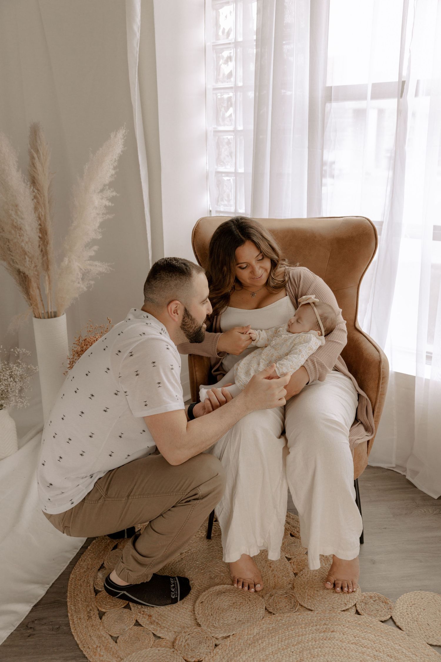 A couple sits together in a tan leather armchair by a bright window with pampas grass decor next to them.