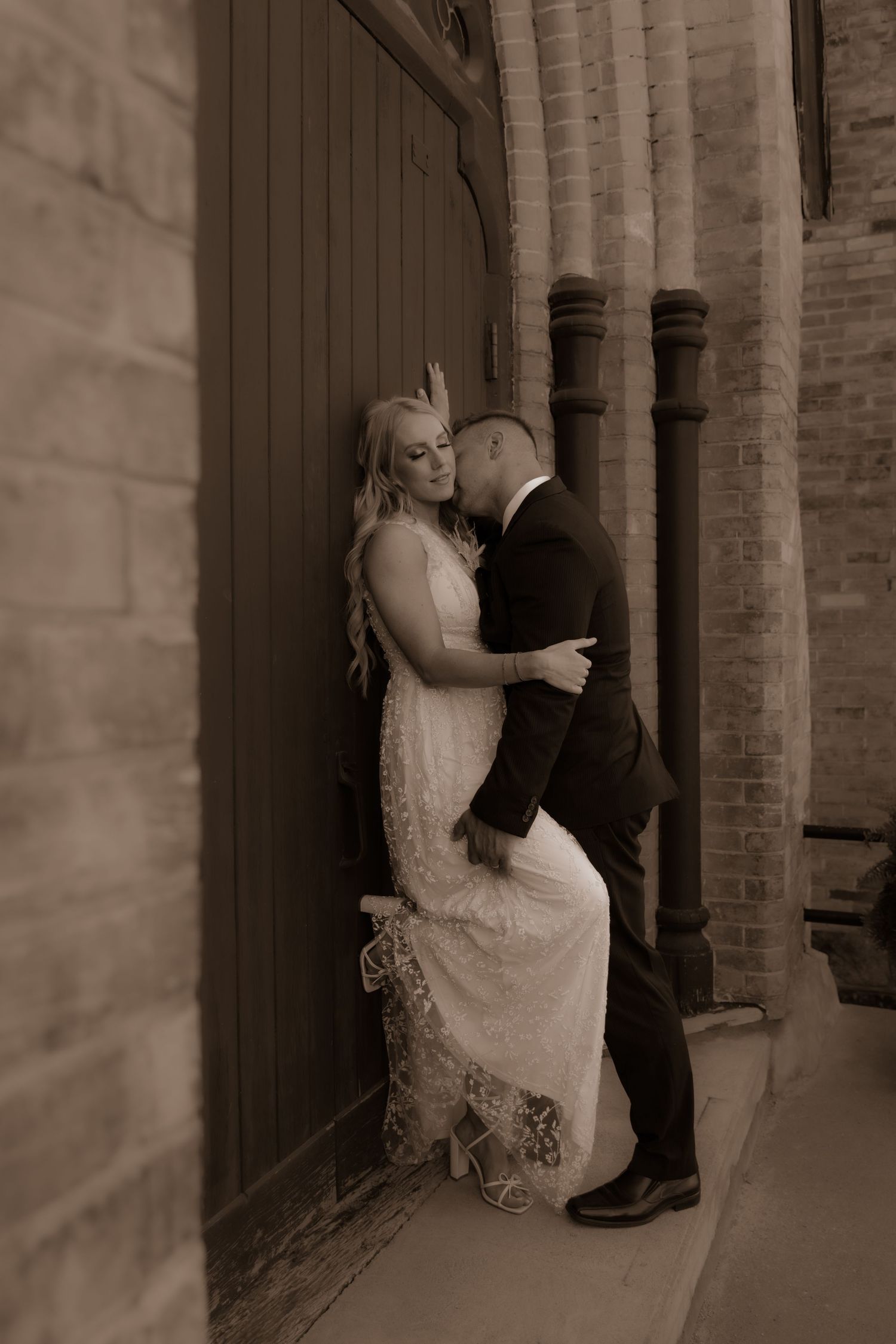 An intimate moment captured in sepia tones against a brick wall.