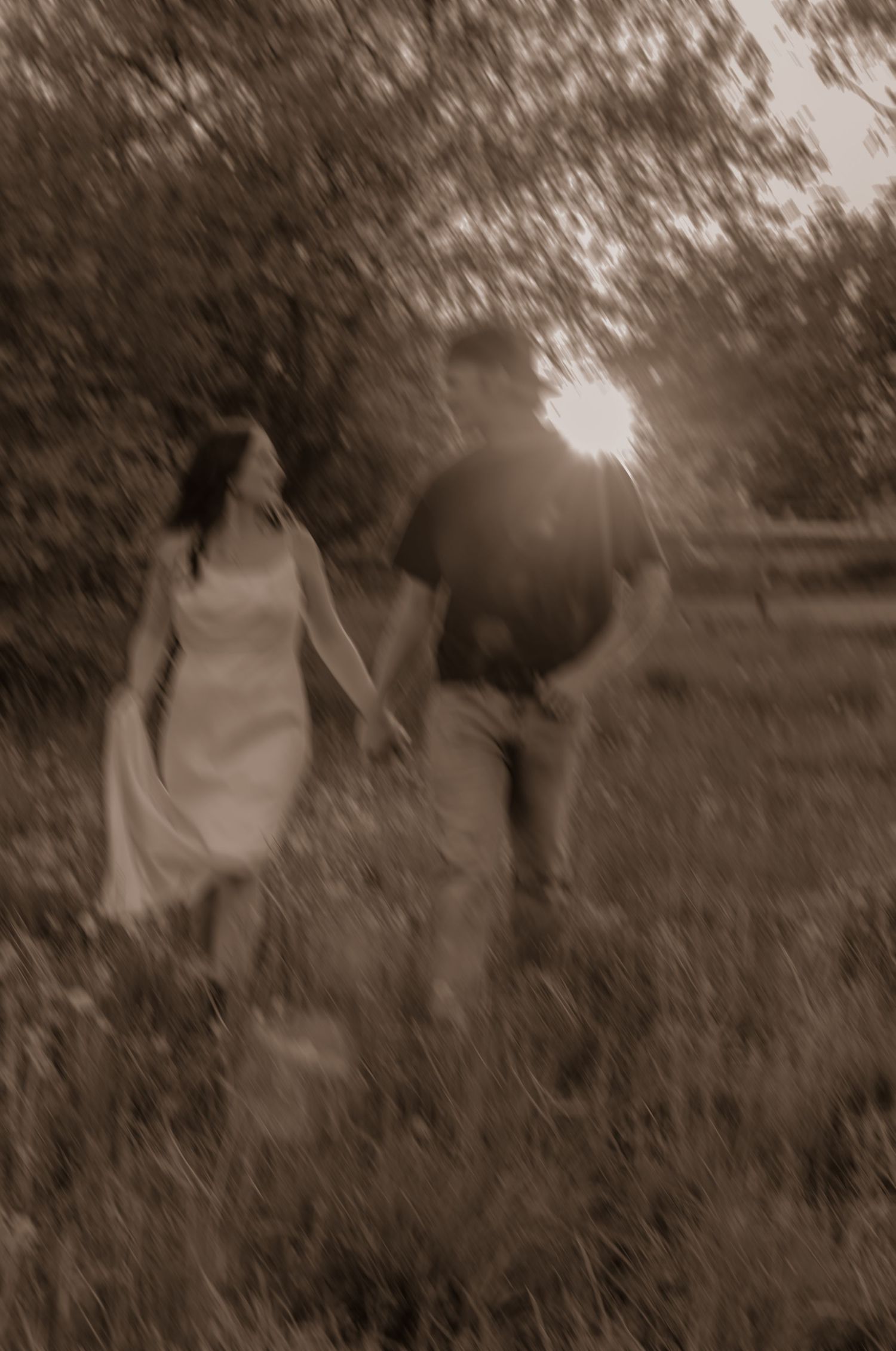 A sepia-toned romantic photograph of a couple holding hands while walking through a sunlit garden path.