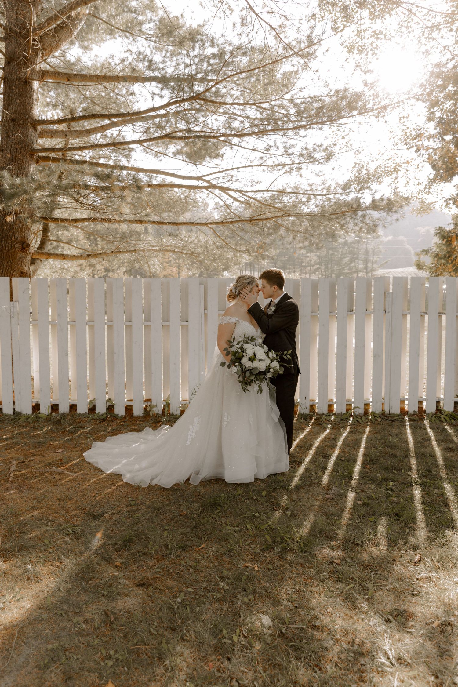 Newlyweds share a tender moment by a white picket fence as sunlight creates dramatic shadows through pine branches.