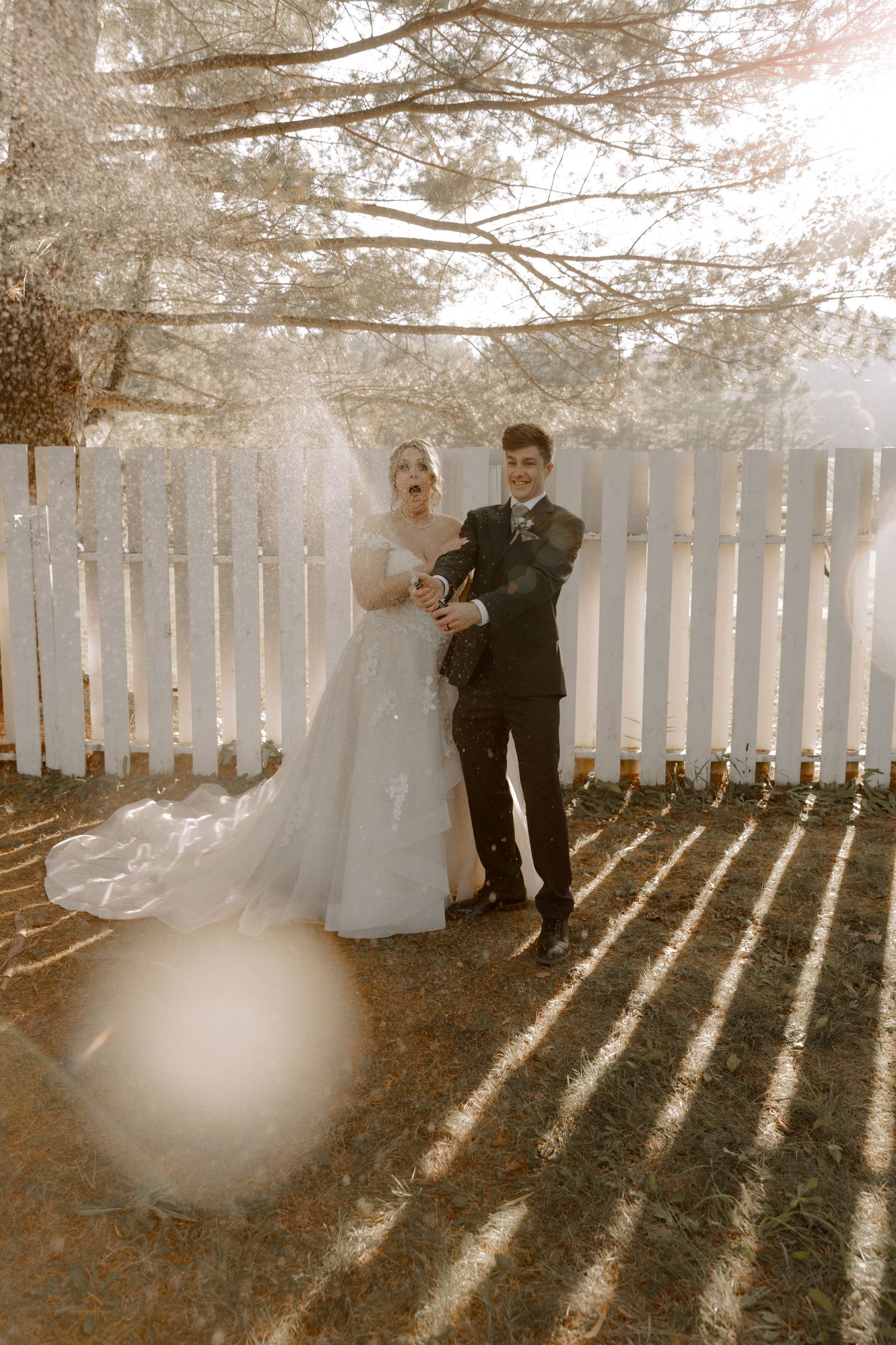 A sun-drenched wedding portrait against a white picket fence with pine trees creating dramatic lighting effects.