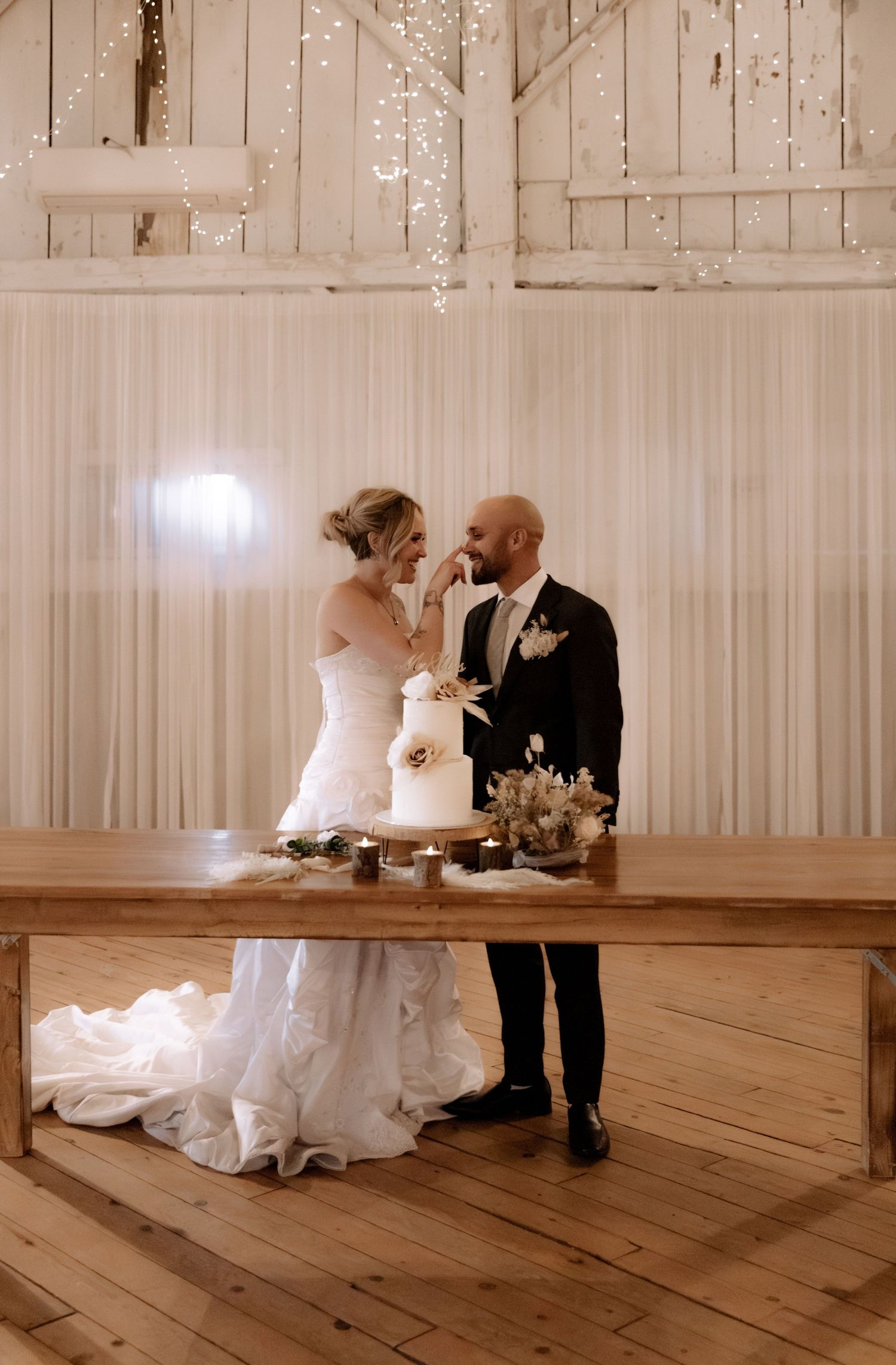Wedding couple stands together by their elegant white tiered cake in a bright rustic venue.