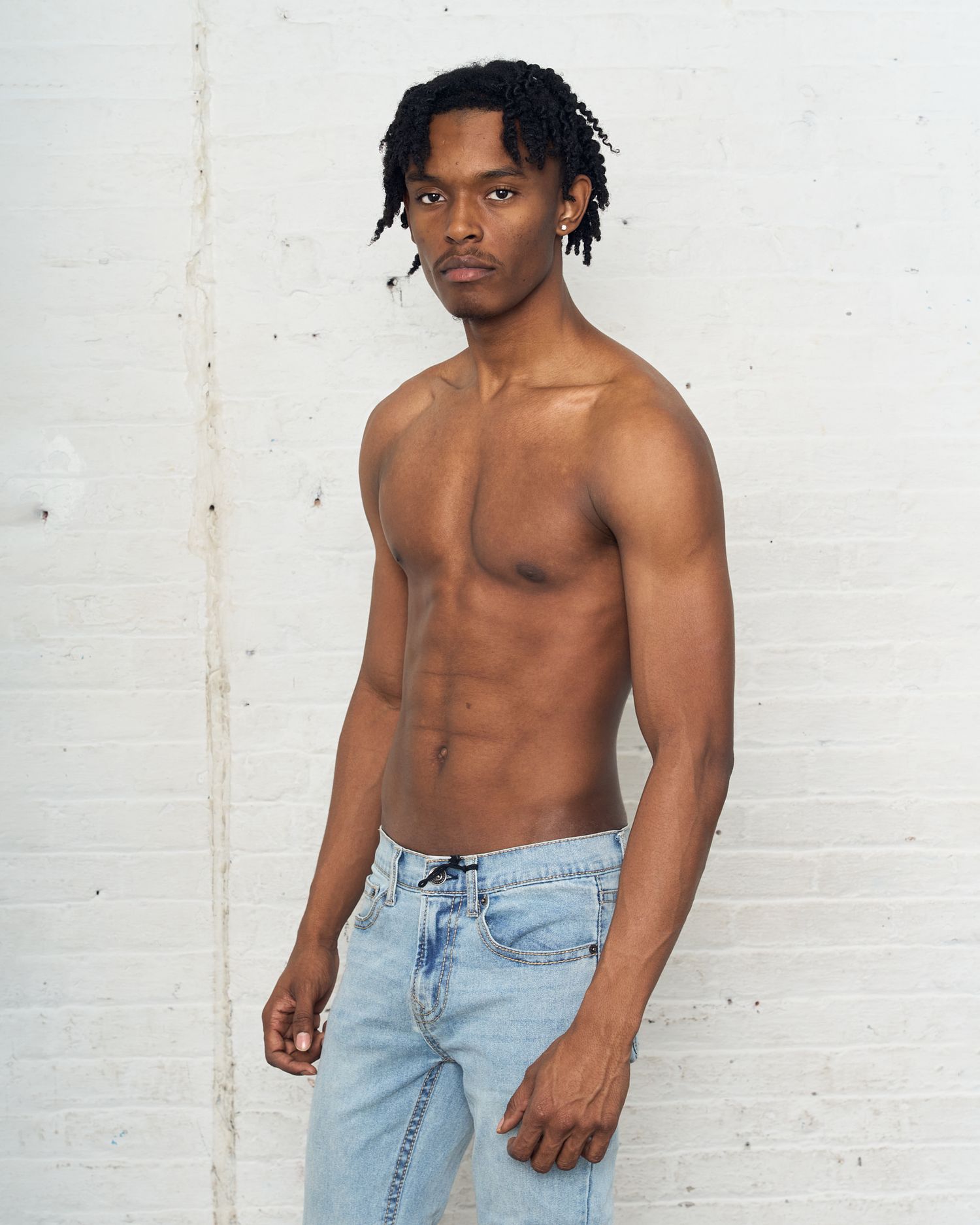Athletic model in light denim jeans poses shirtless against white brick wall. Modeling Digitals