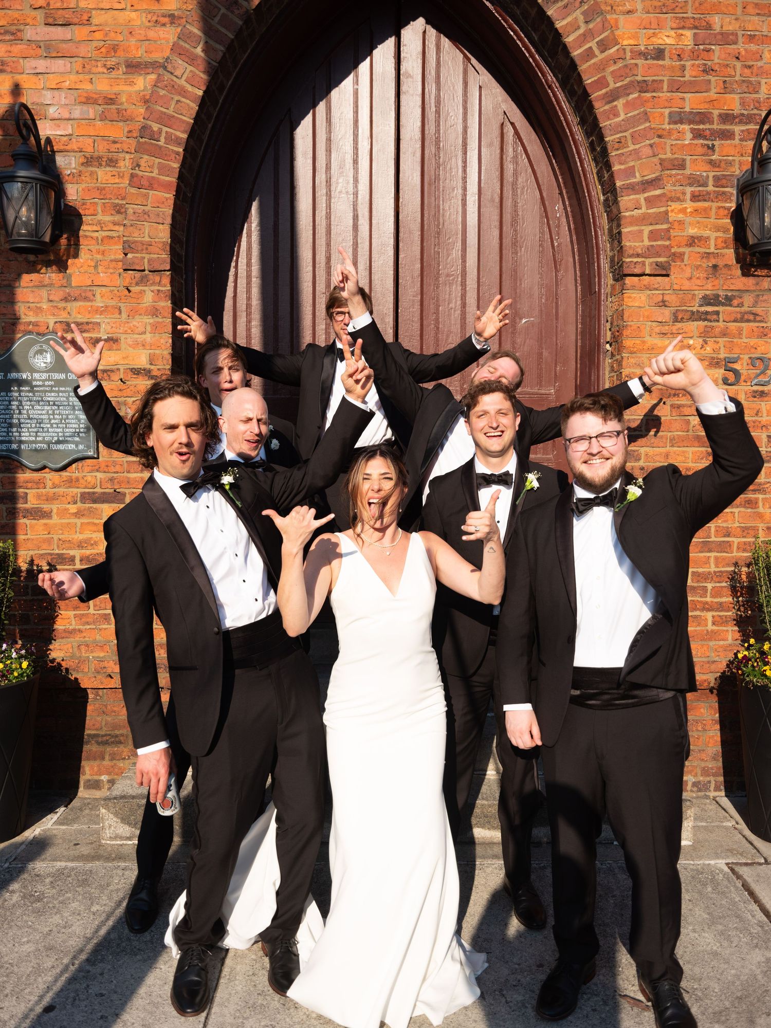 A wedding party in formal attire poses playfully in front of an arched brick church doorway.