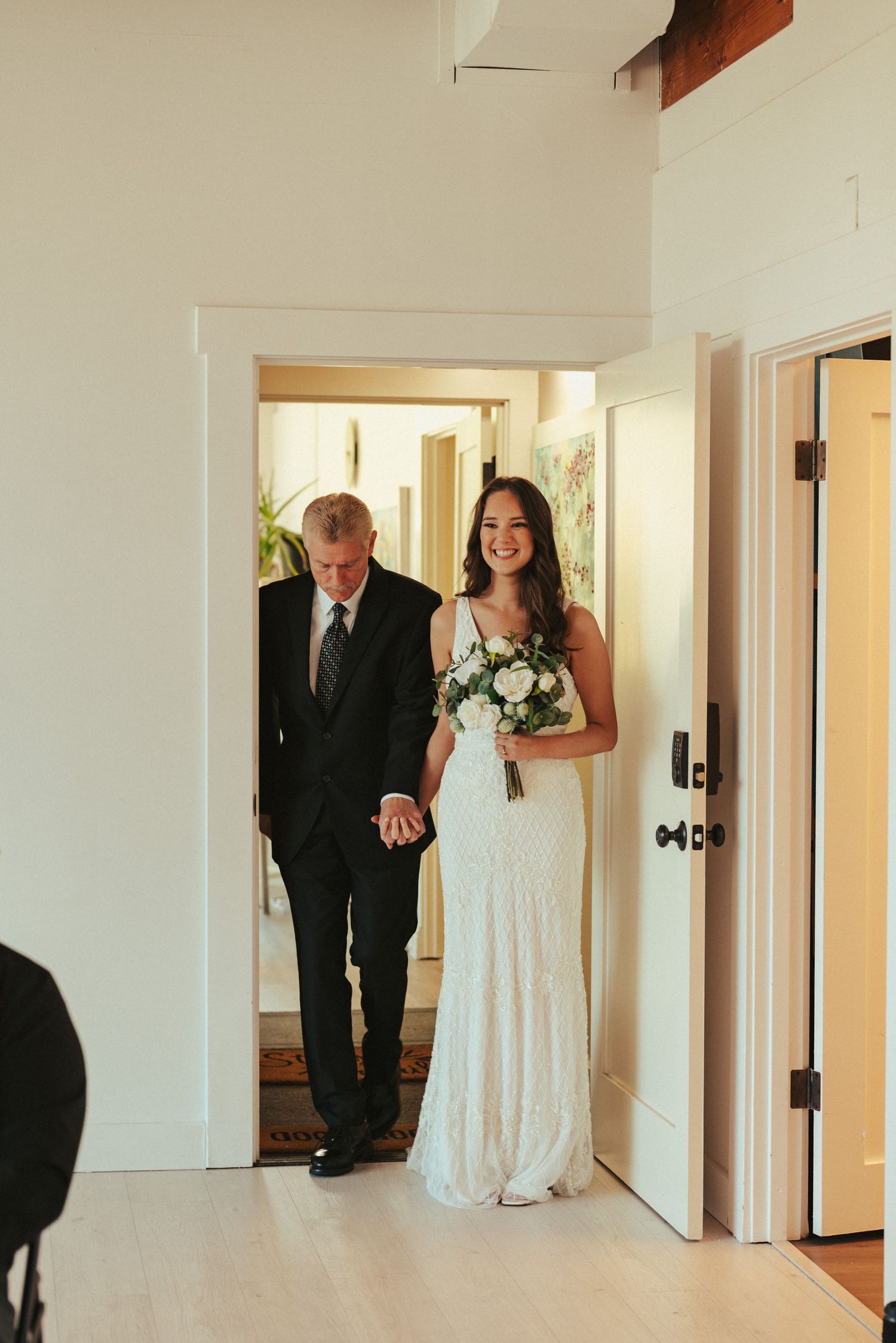 A bride in white holding a bouquet enters through a doorway with her escort.