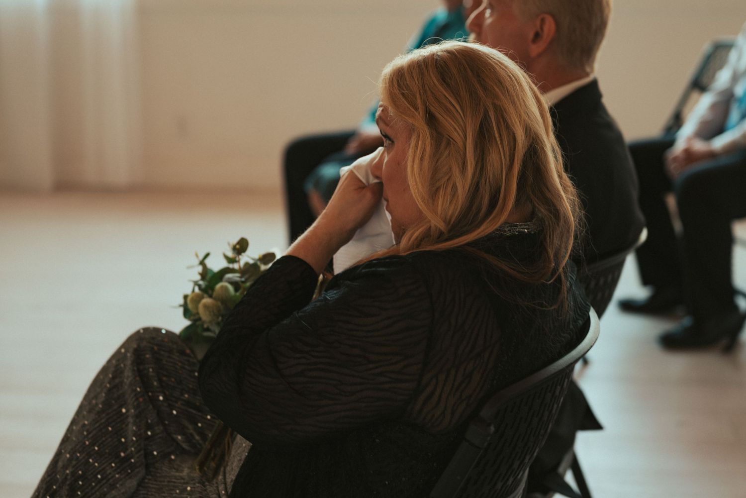 Three guests sit in chairs at a wedding ceremony in a modern indoor venue.
