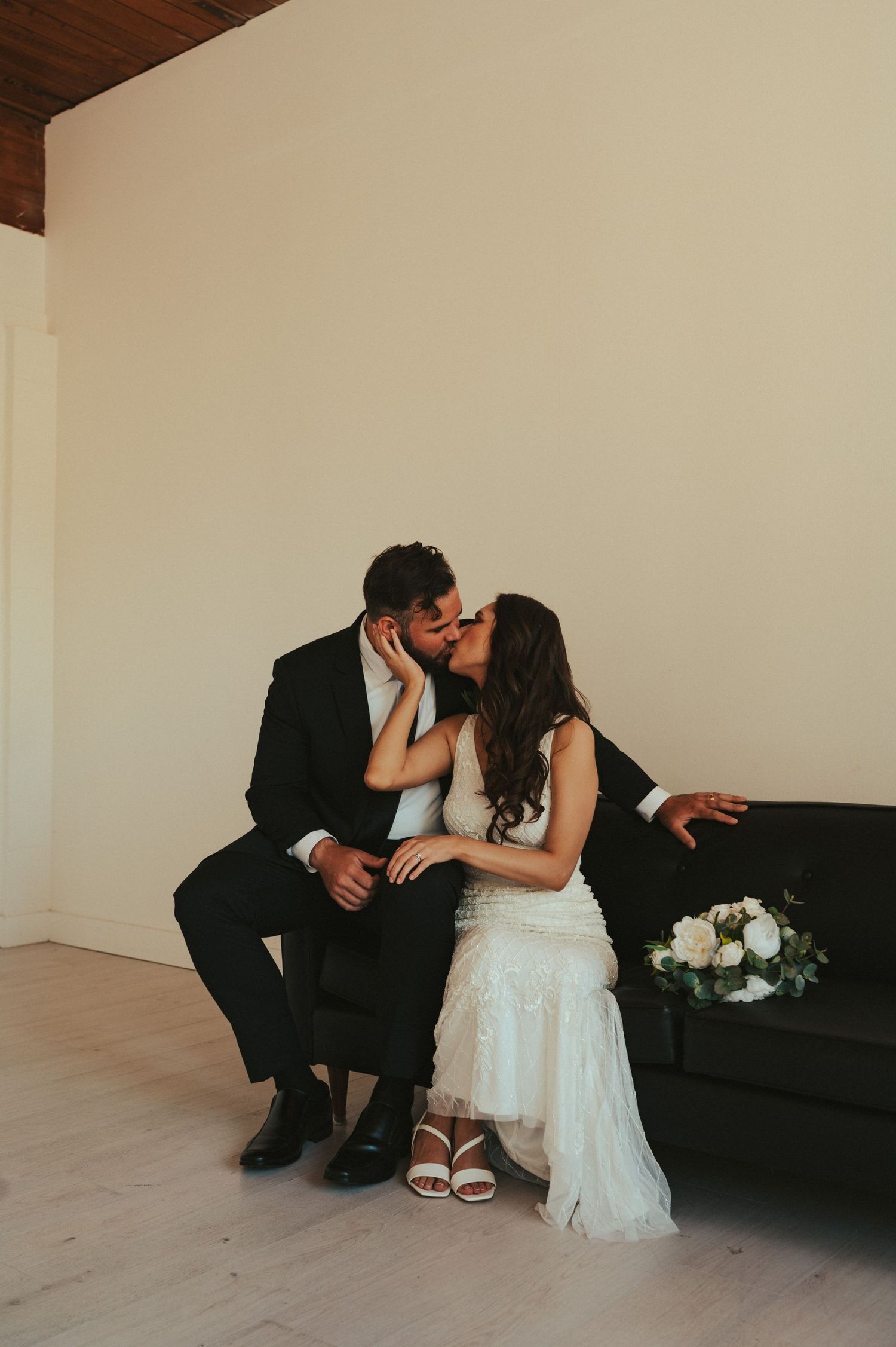 A tender moment between newlyweds sitting on a dark couch in a minimalist room.