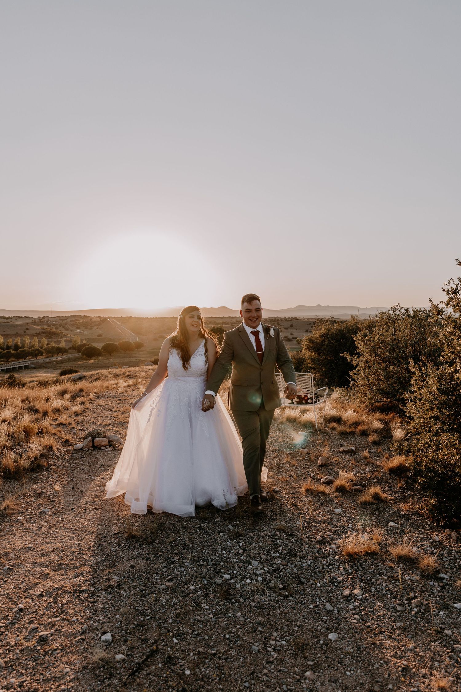 Newlyweds walk together on rocky desert path with setting sun creating warm golden light.