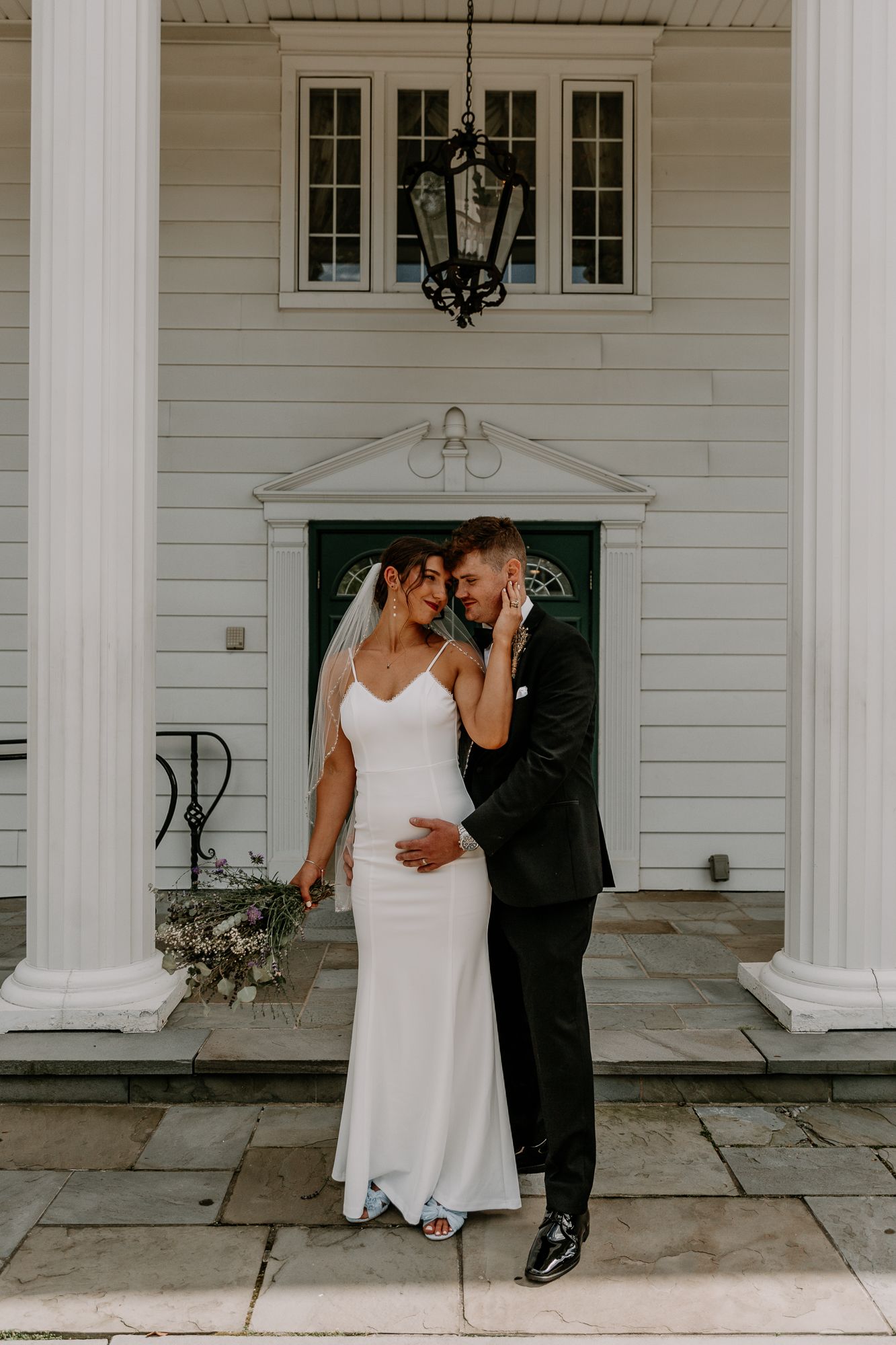 A couple shares an intimate moment on a white columned porch of a classic building.