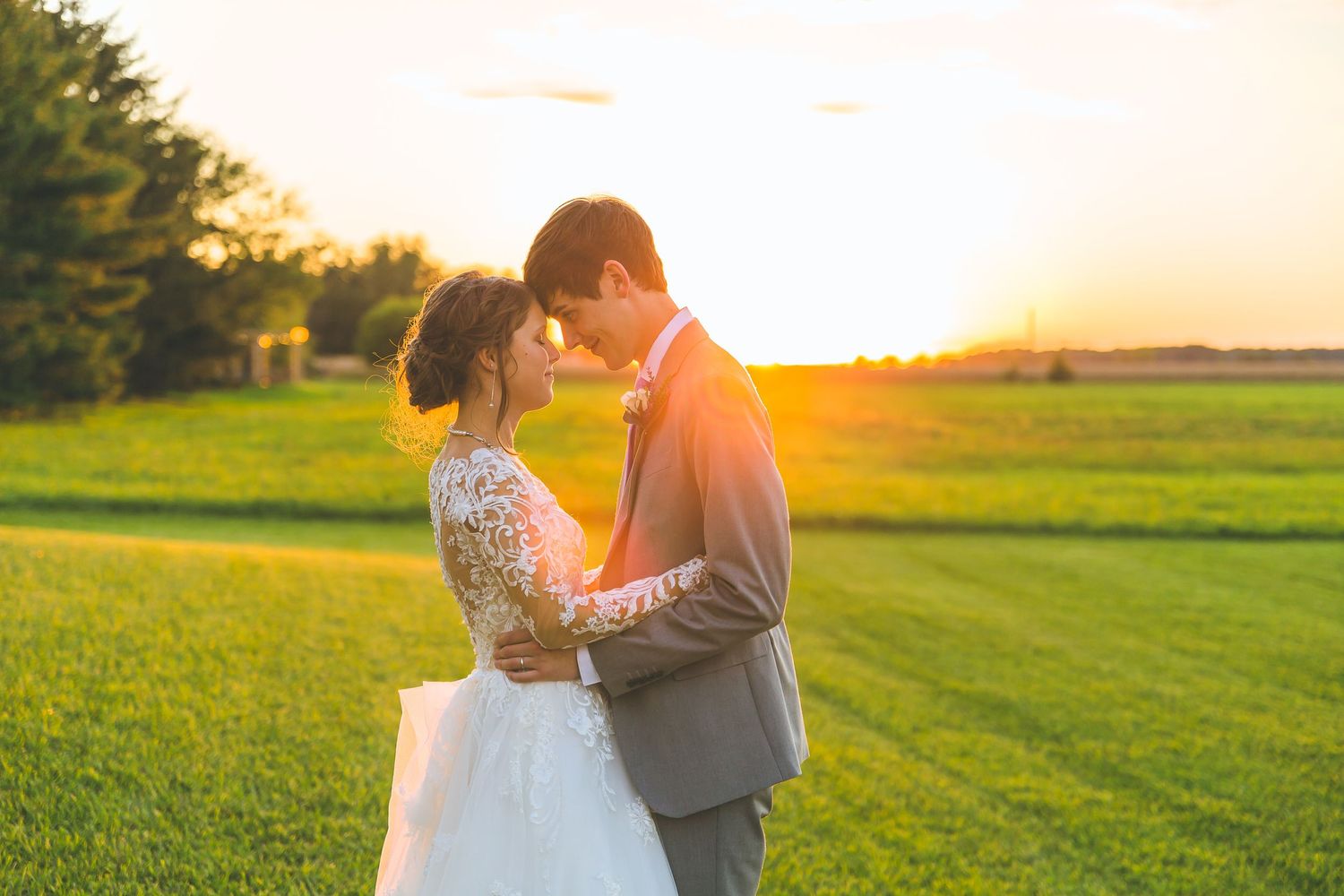 A couple embraces on a grassy field during golden hour with sunlight streaming through.