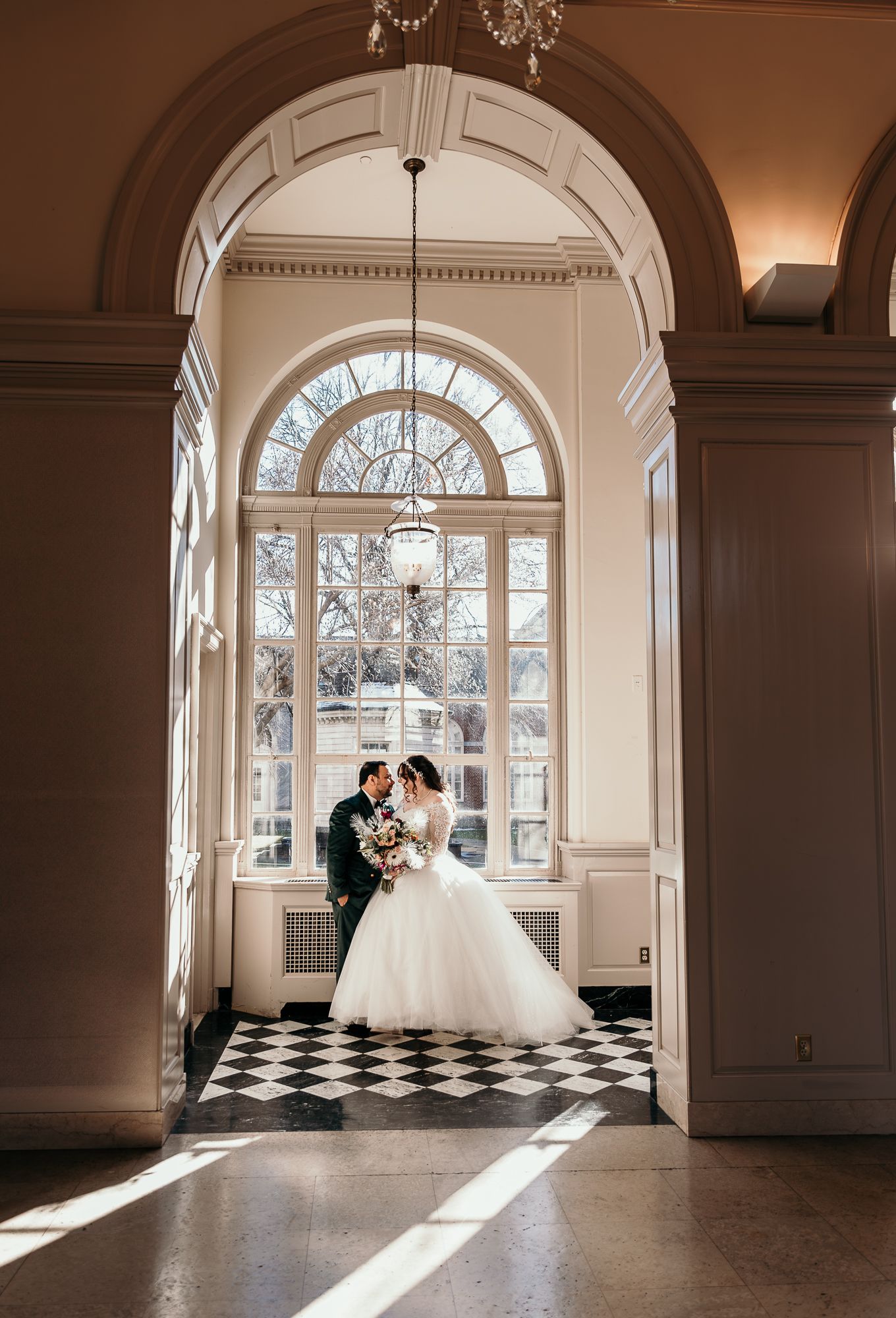 A romantic wedding portrait under an arched window with checkered floors and streaming natural light.