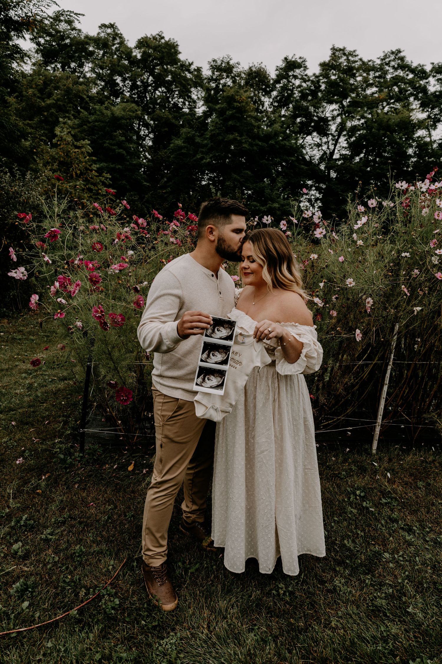 A couple embraces in a flower garden while looking at an ultrasound photo.