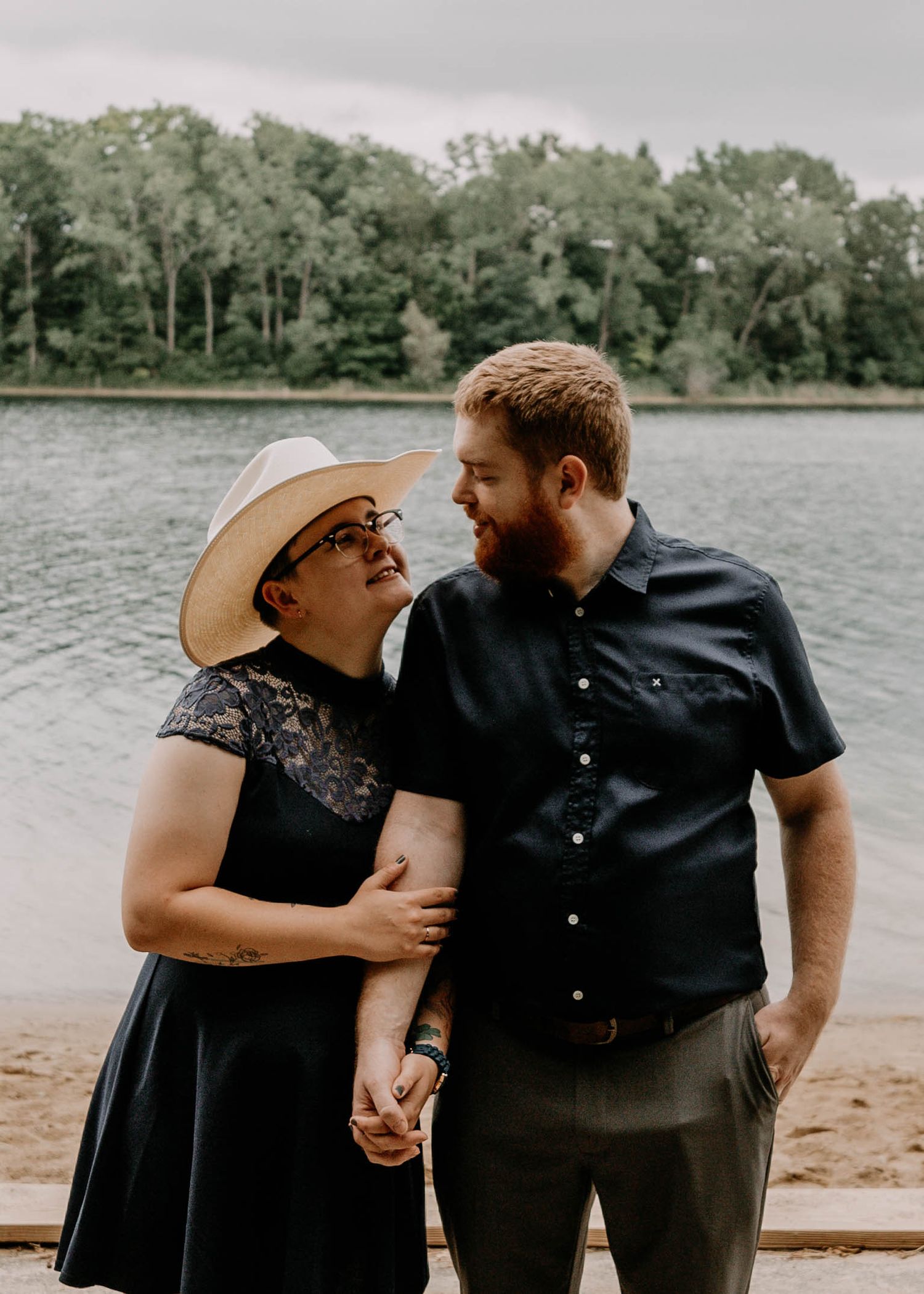 Two people sharing an intimate moment at the edge of a lake with trees in the background.
