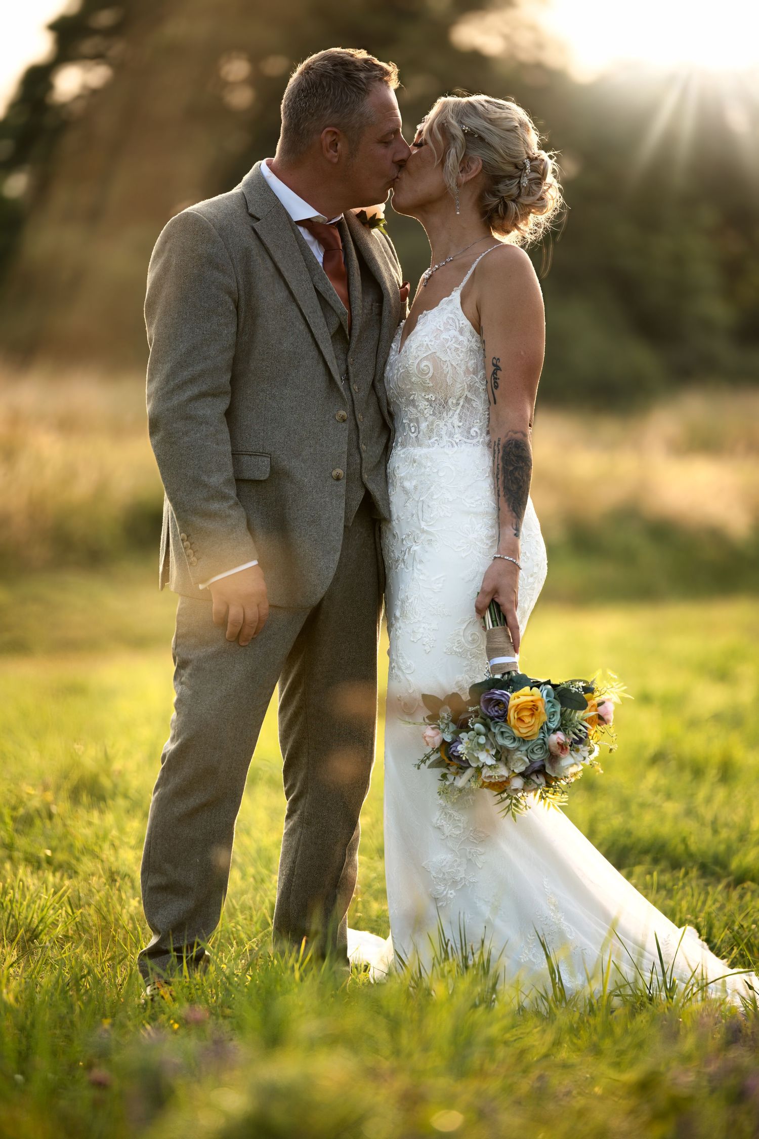 Newlyweds share a kiss in a sunlit field while the bride holds a vibrant bouquet.